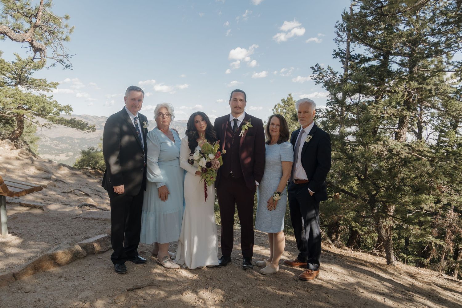 Wedding party family portrait of six people standing together among pine trees with mountain landscape behind them.