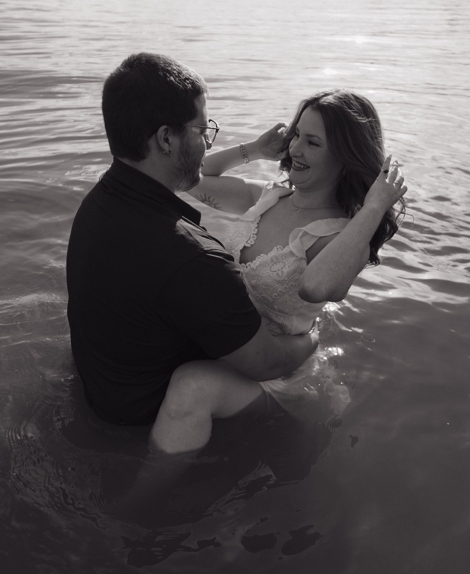 Black and white romantic photo of couple embracing in water, woman in white dress sitting on mans lap.