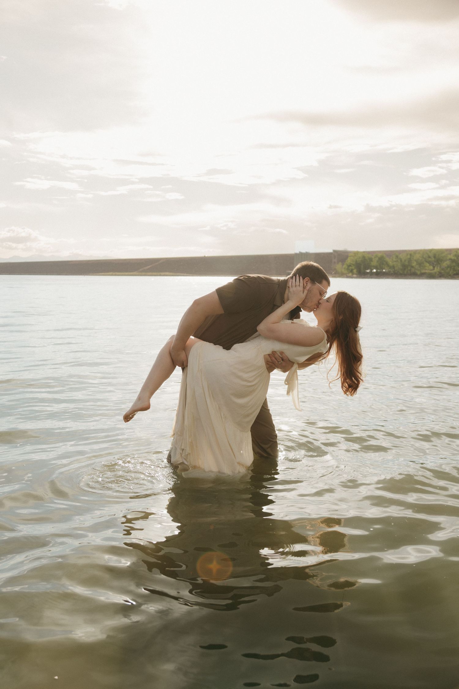 A romantic couple kissing in a lake at sunset, man carrying woman in white dress in golden light.