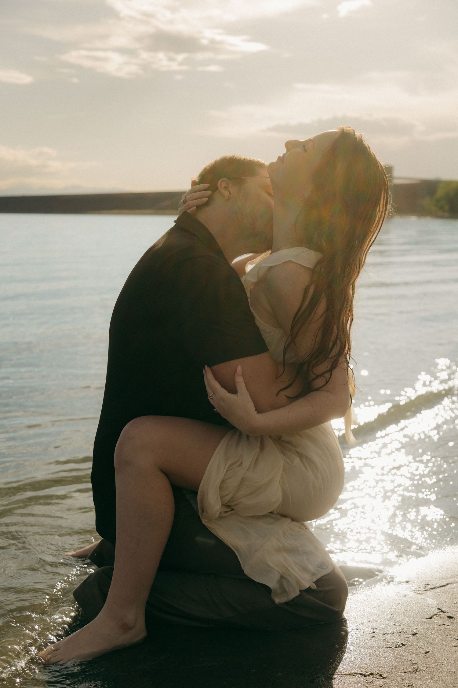 Romantic couple embracing and kissing on sandy beach shore with warm glowing sunset light.