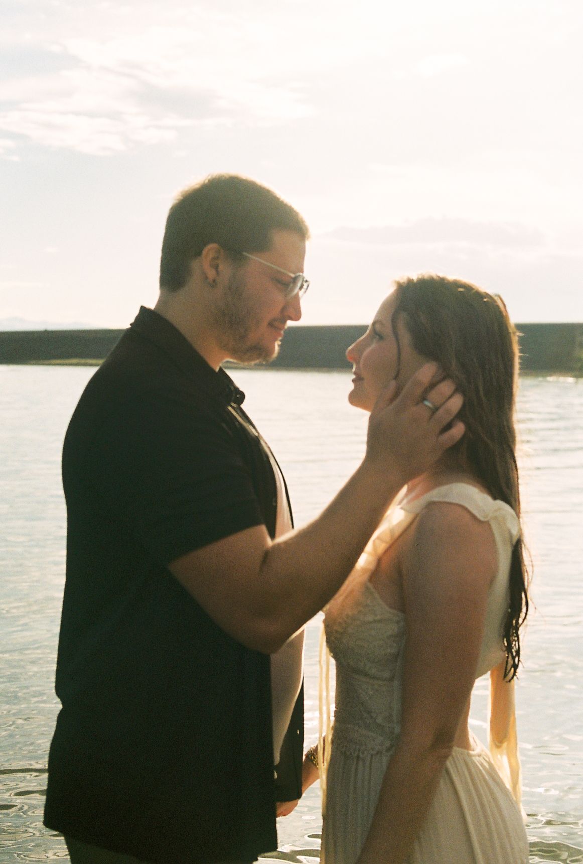 Couple in a tender embrace by the water at golden hour, man gently cupping womans face in warm backlit sunlight.