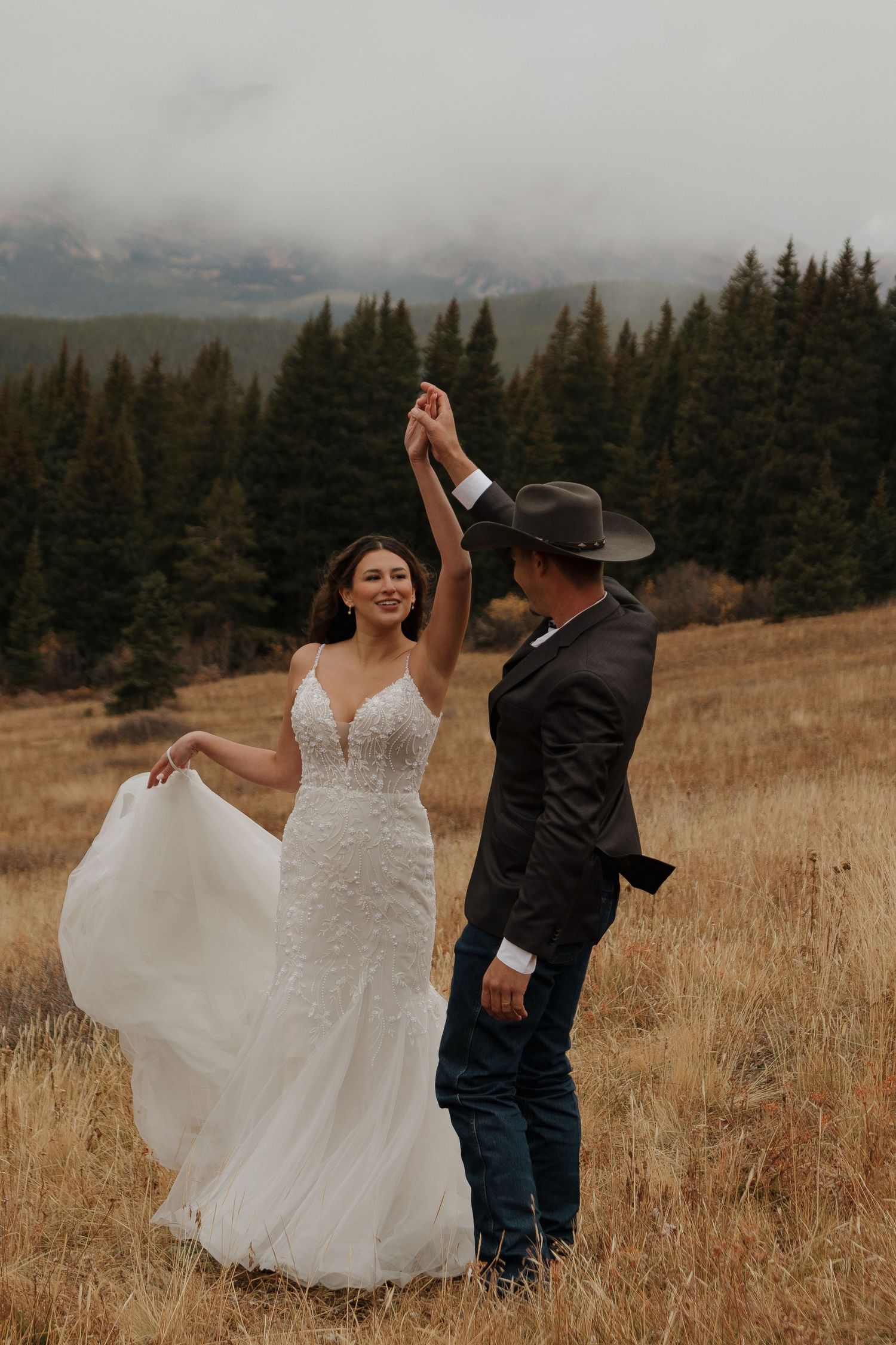 Bride in lace gown dances with groom in cowboy hat on a mountain meadow at sunset.
