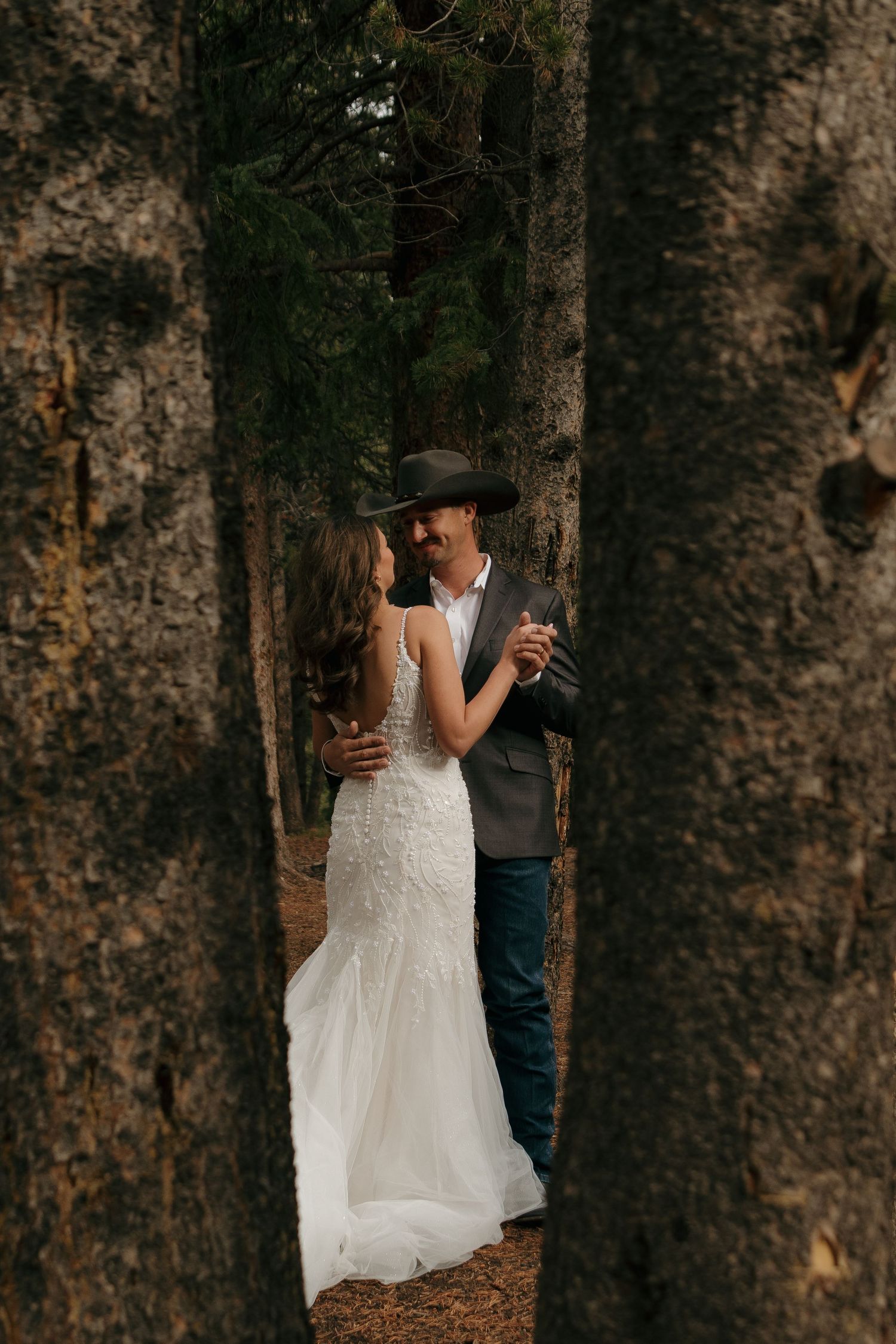 A couple in wedding attire embraces among tall forest trees.