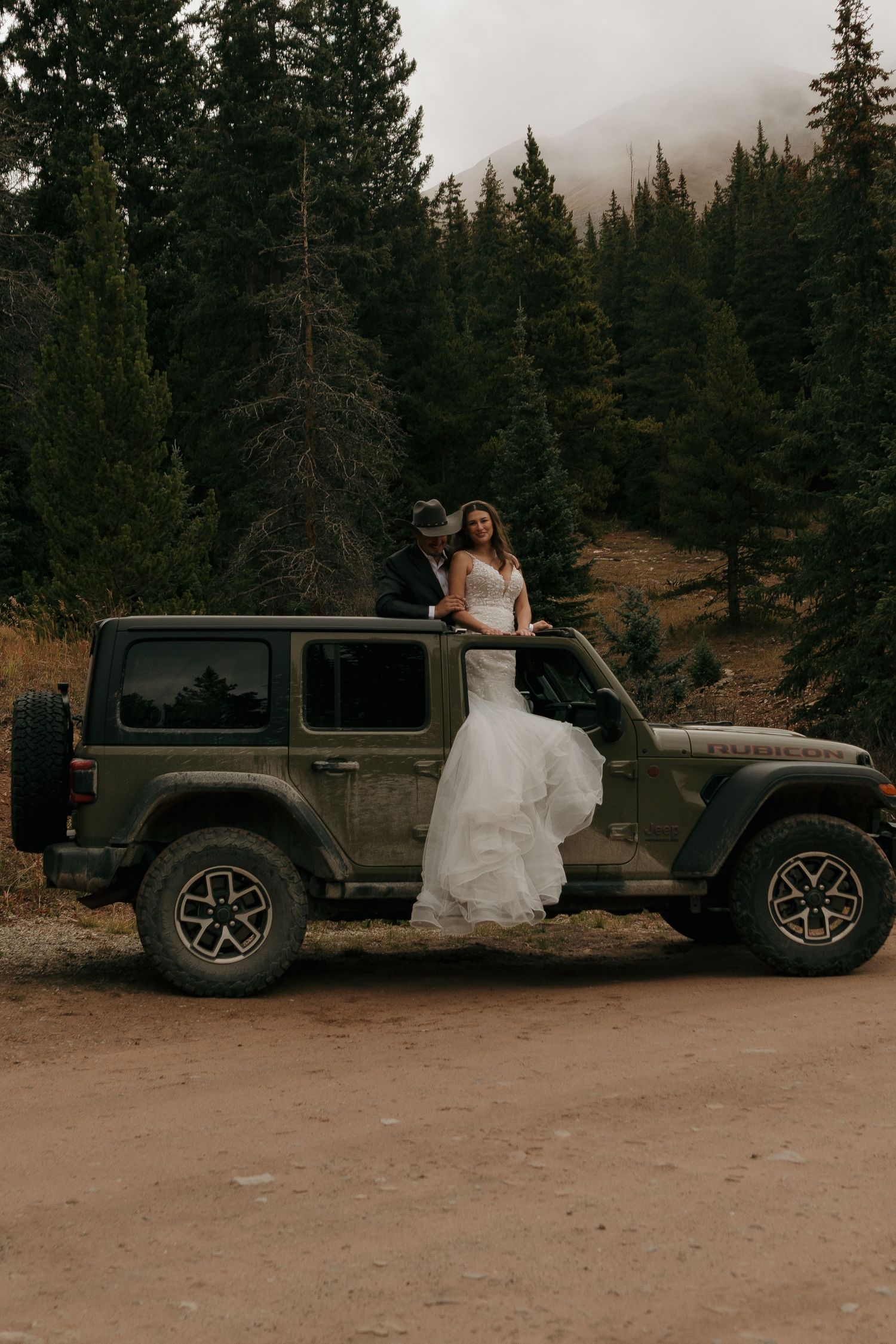 A white wedding dress contrasts against a dark Jeep Wrangler parked in a forest setting.