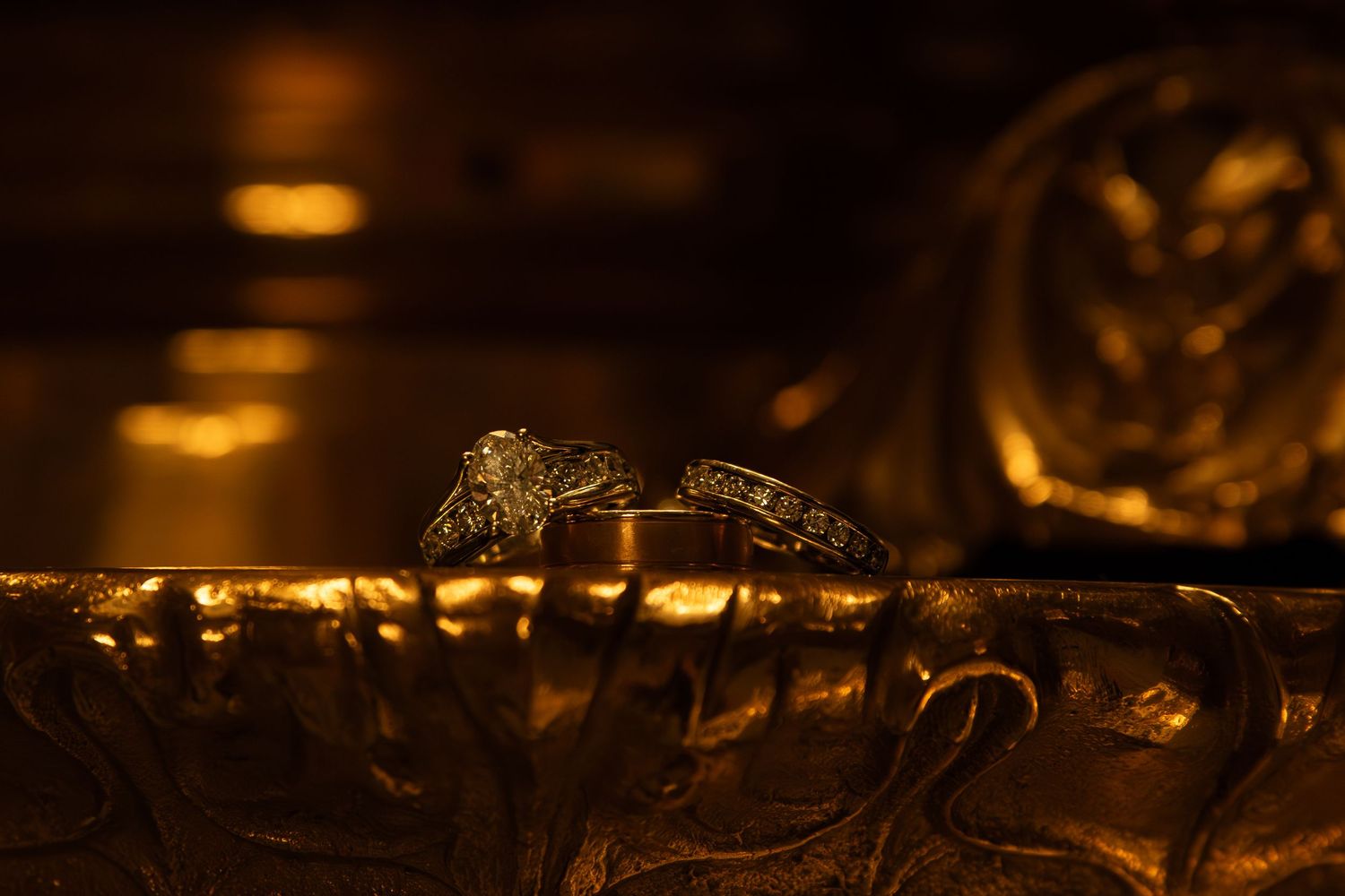 Two ornate wedding rings sit on a reflective golden surface in warm ambient lighting.