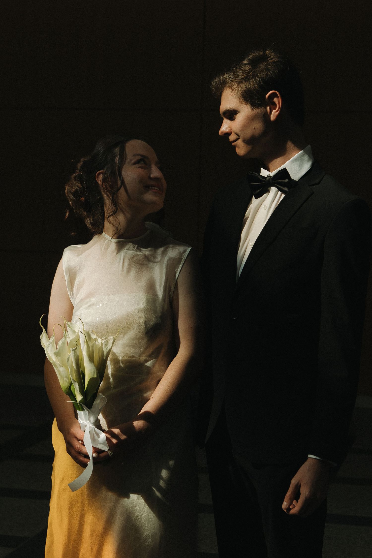 A couple in formal attire stands together in dramatic lighting holding white flowers.