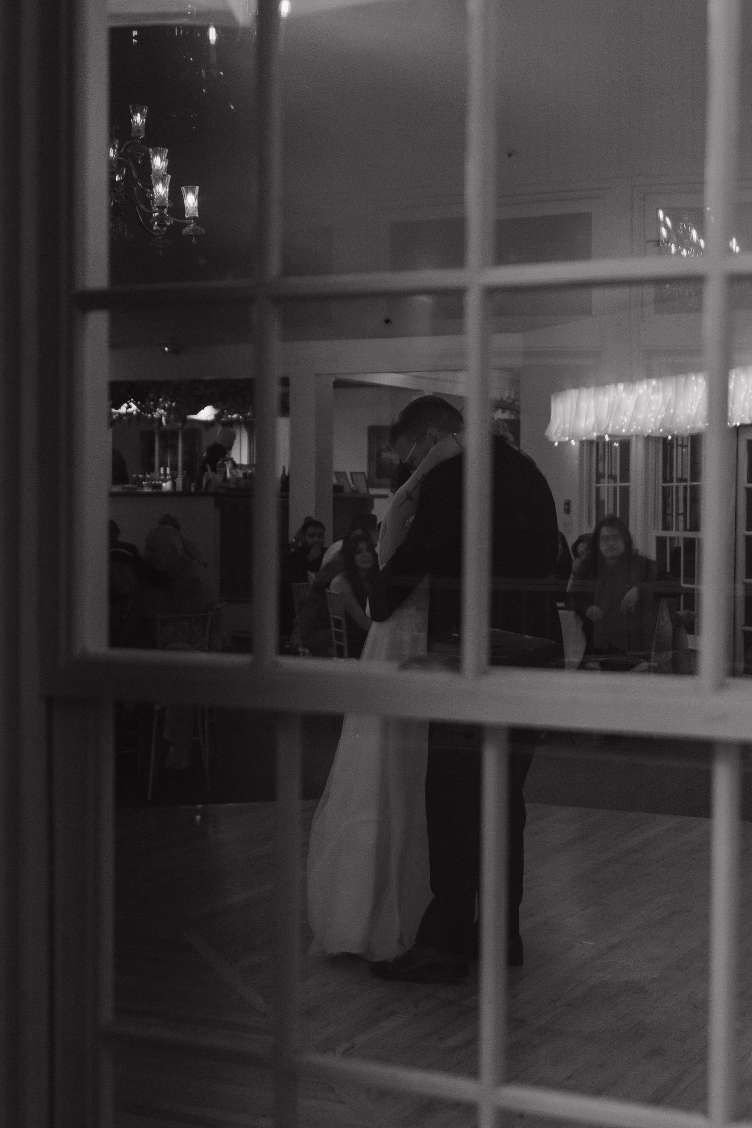 A black and white view through window panes of a couple dancing at an indoor reception venue.
