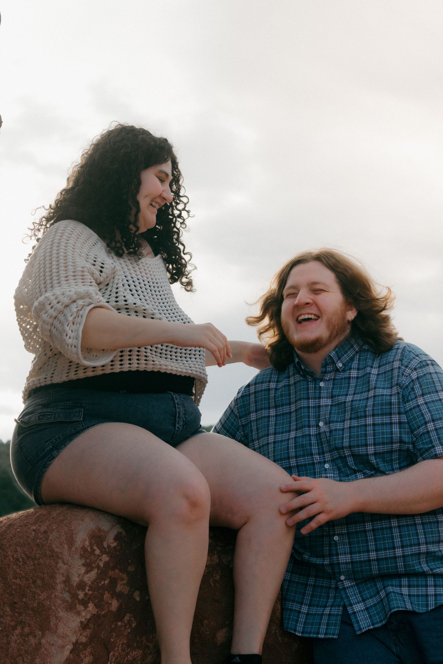 A couple shares genuine laughter while sitting on red rocks outdoors.