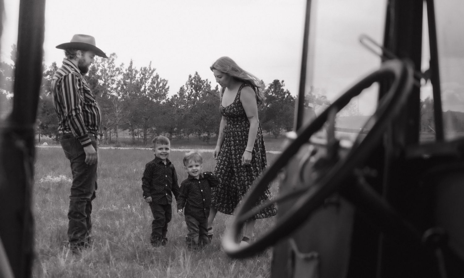 Black and white photo of family gathering in field seen through vintage truck window.
