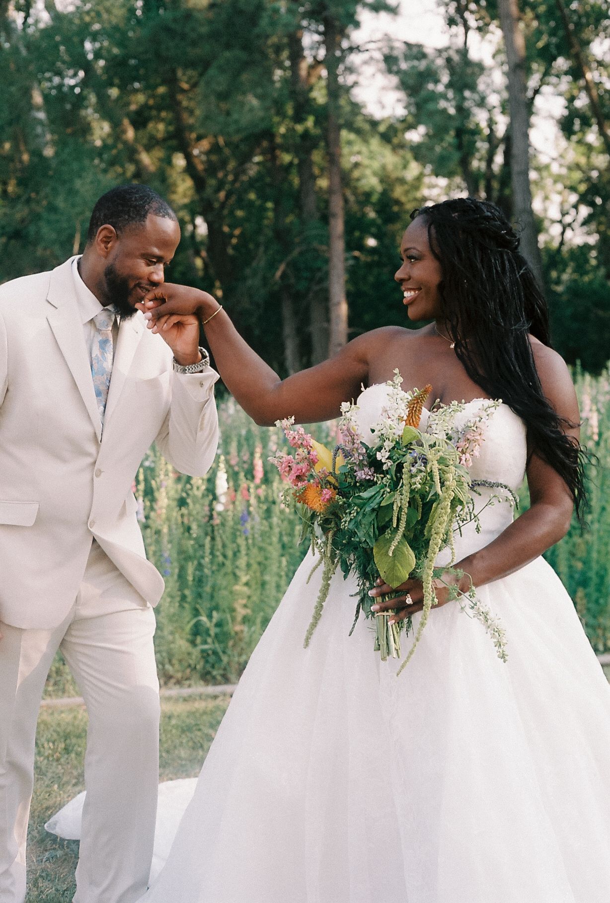 Groom in beige suit kisses bride's hand as she smiles holding colorful bouquet outdoors at wedding.
