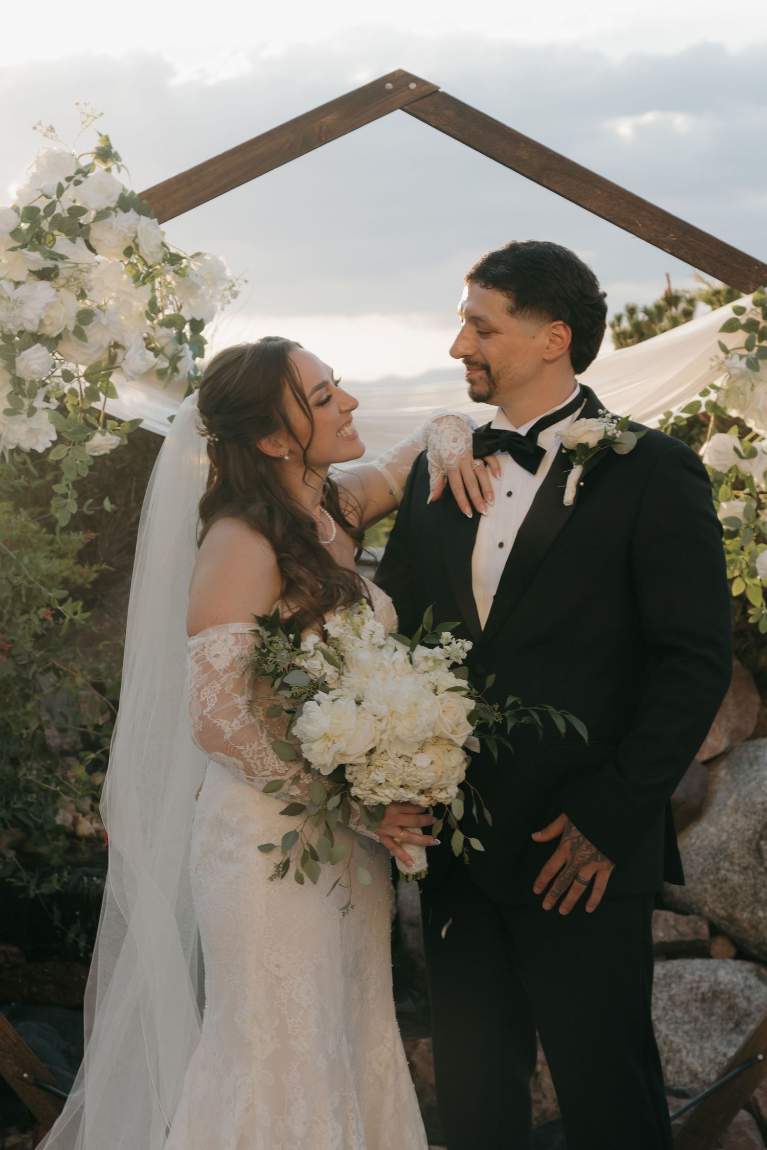 Bride and groom posing under wooden arch with white floral arrangements at outdoor wedding ceremony.