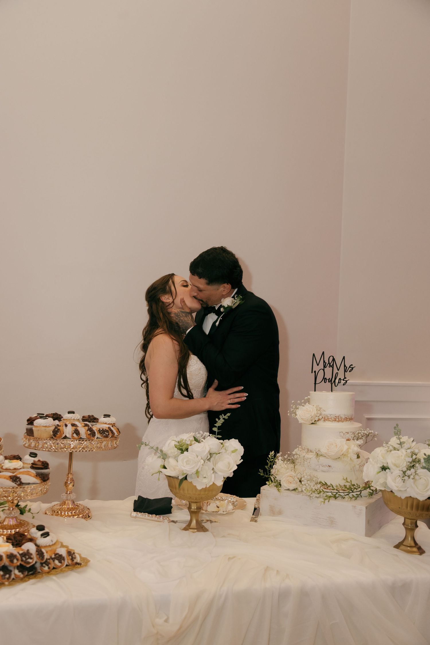 Newlyweds share a kiss at a dessert table with a white wedding cake and floral arrangements.