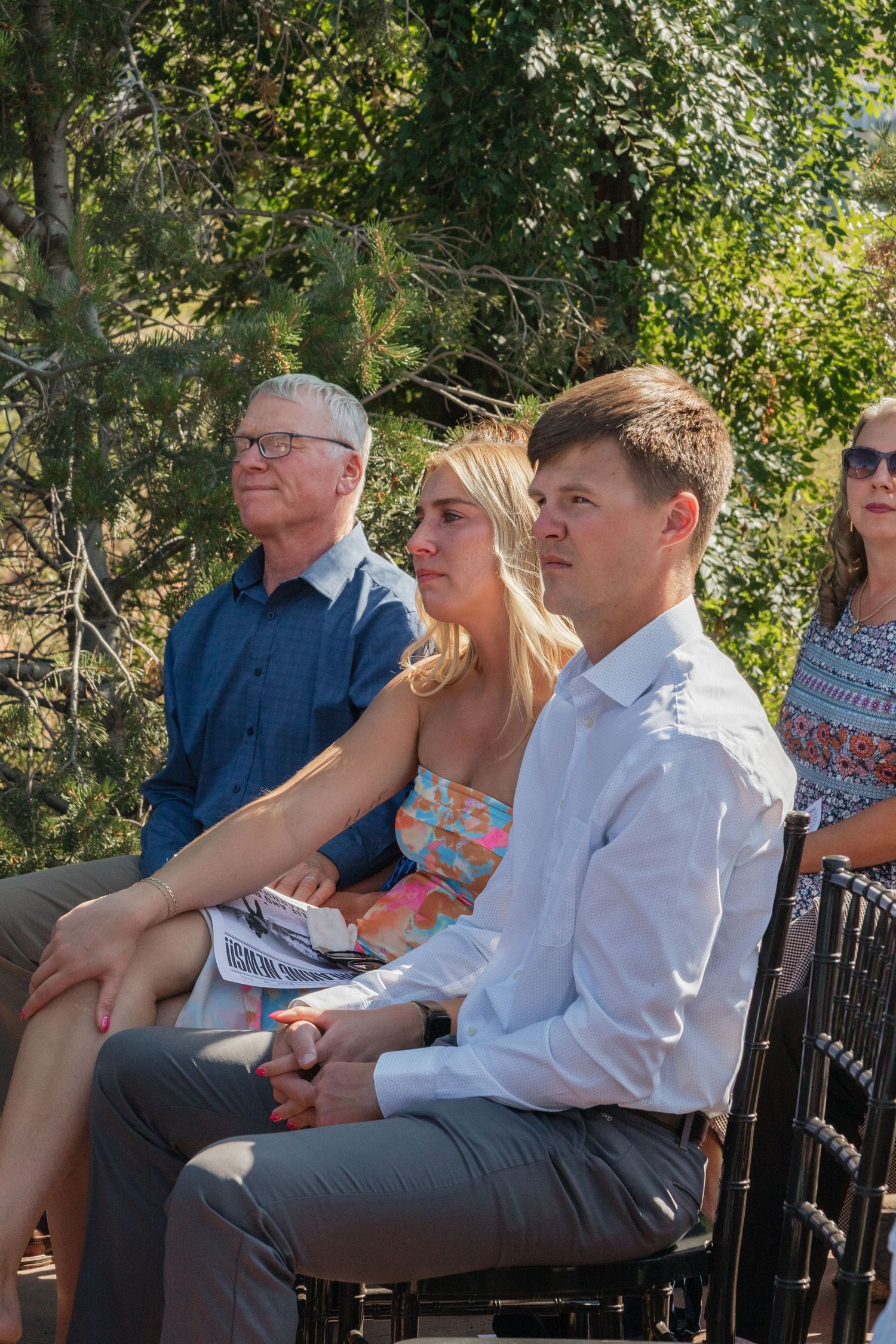 Wedding guests seated outdoors watching ceremony under natural tree canopy.
