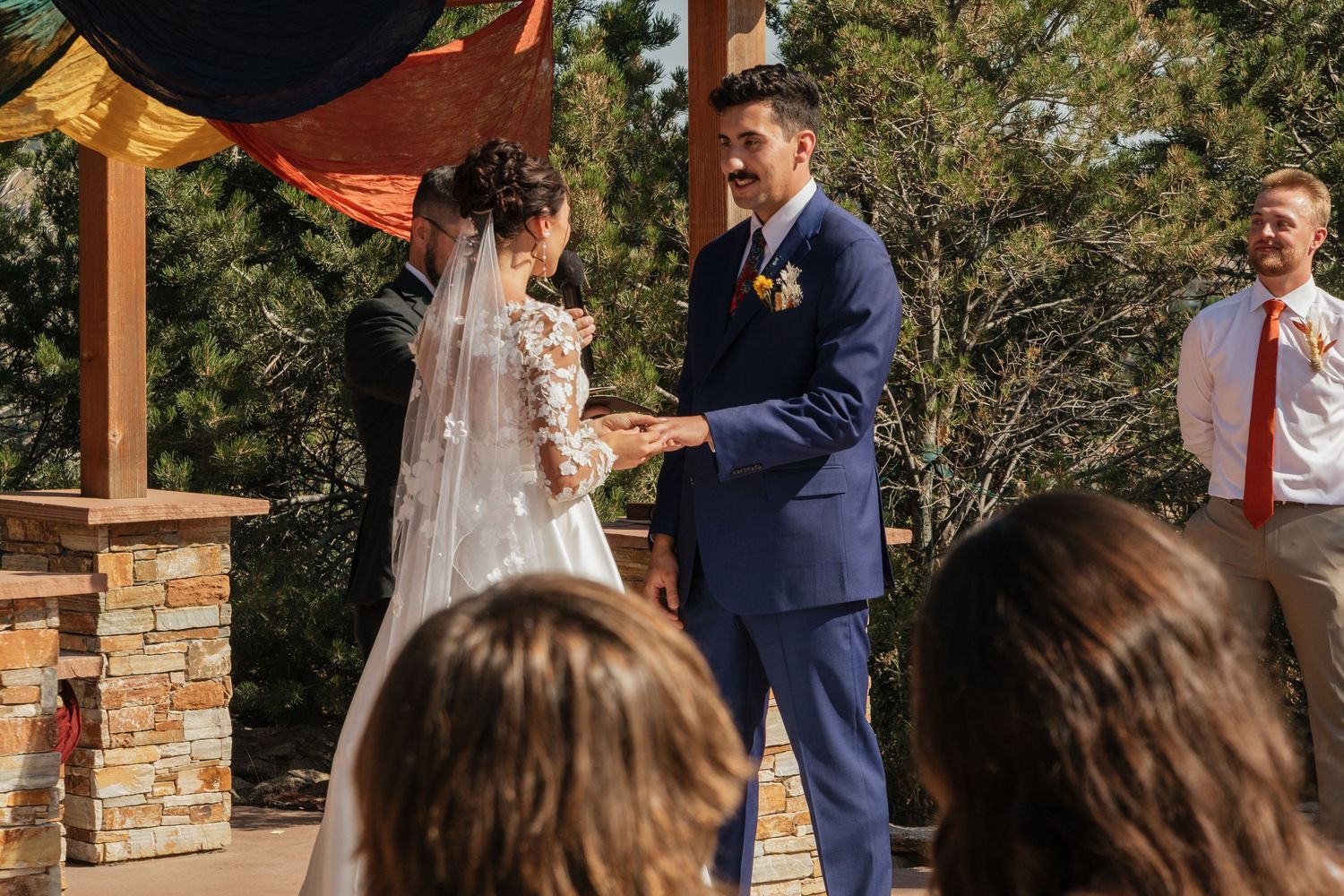 A wedding ceremony taking place under a wooden pergola with guests watching the proceedings.