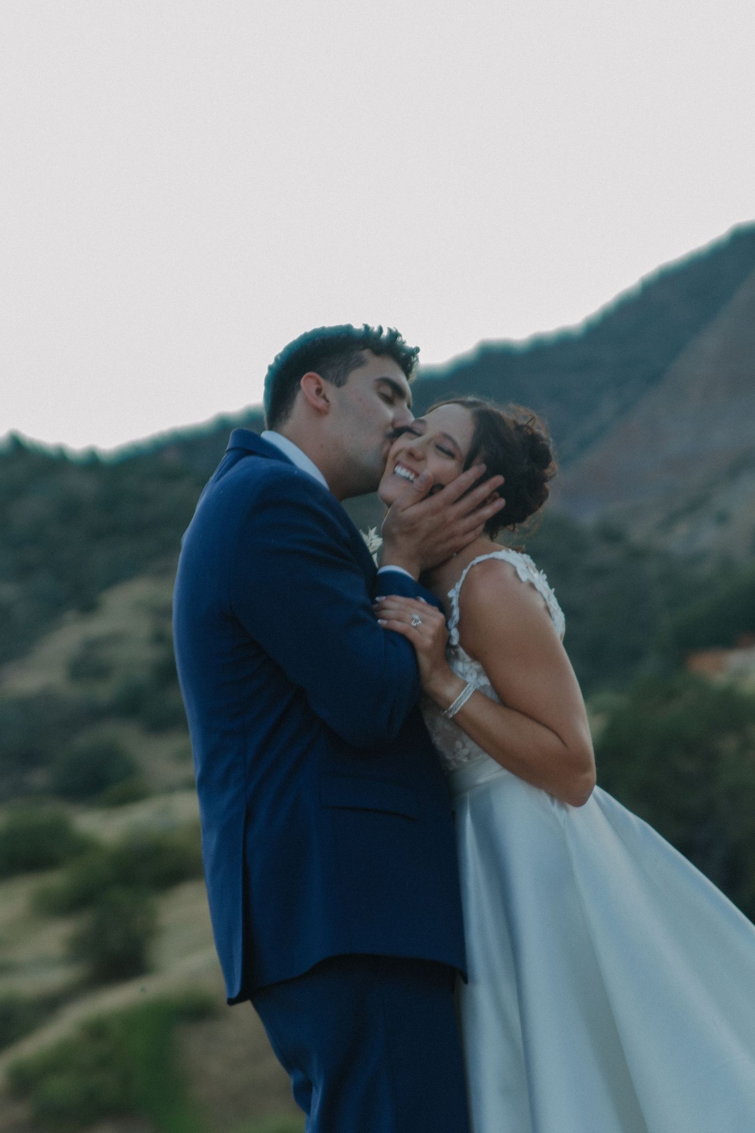 A romantic wedding couple sharing intimate kisses against a mountain backdrop at sunset.