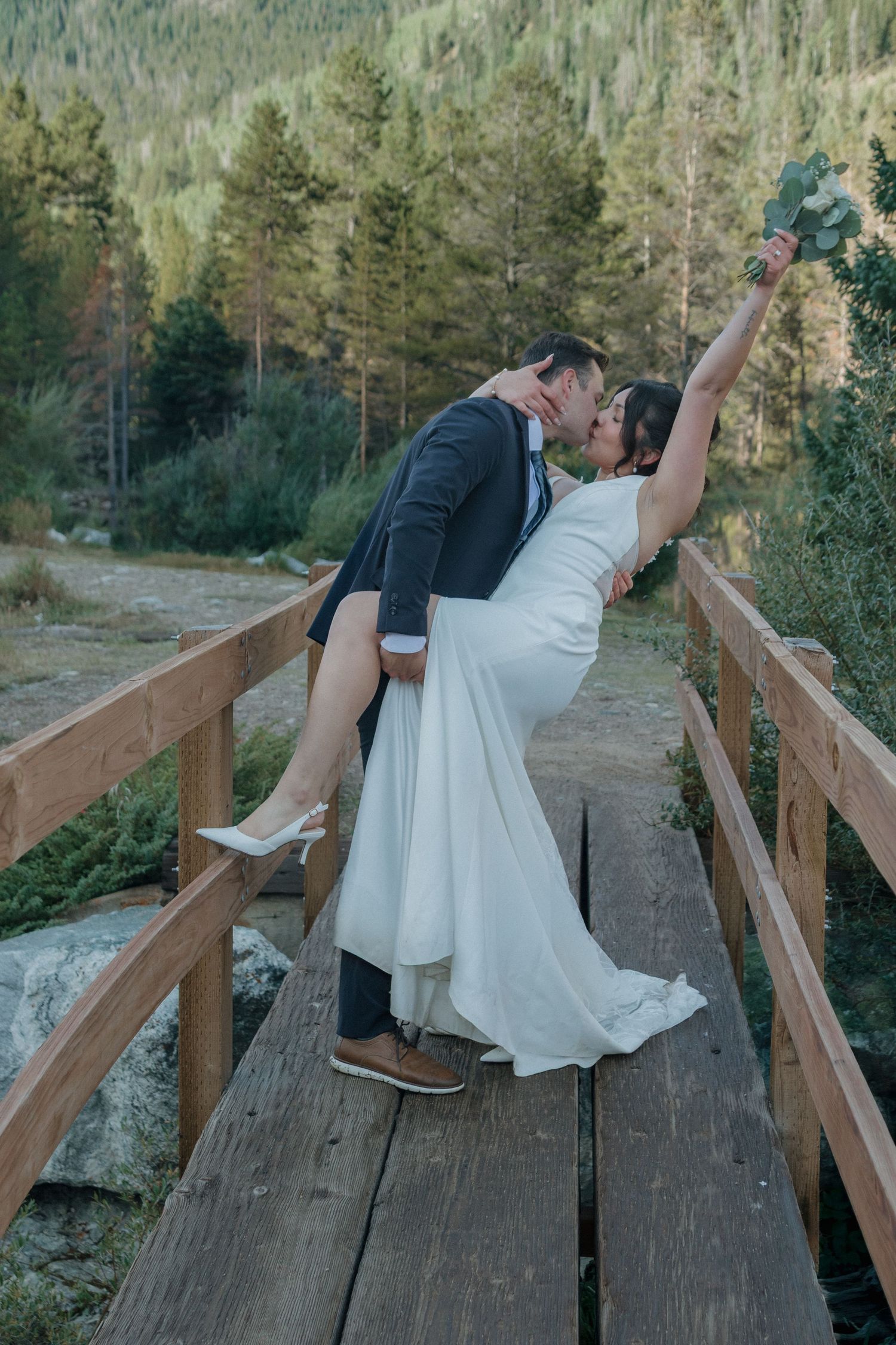 Romantic dip kiss on wooden bridge with mountain wilderness backdrop.