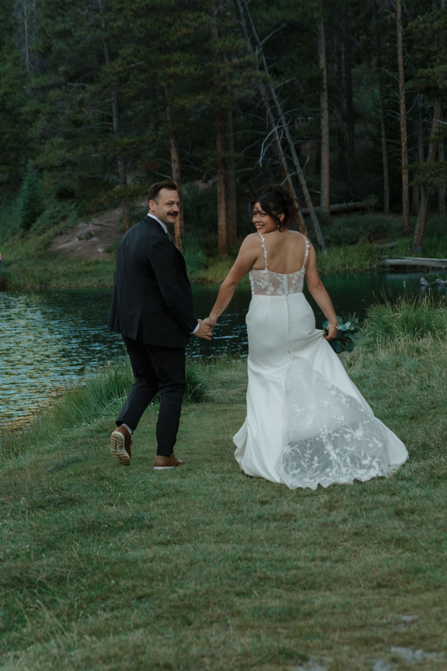 Couple walks hand in hand along grassy lakeshore during their mountain wedding.