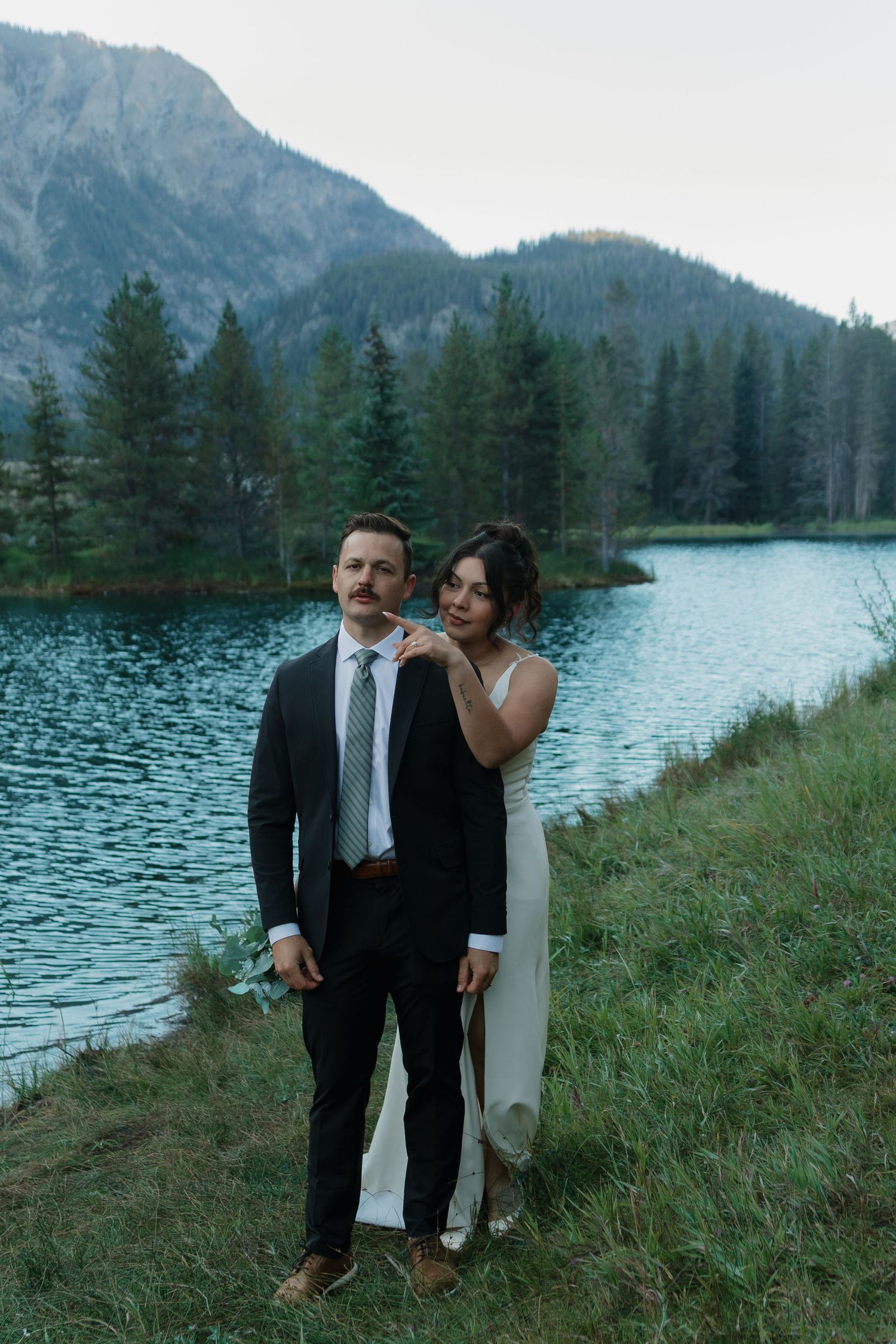 A couple shares romantic moments by a serene mountain lake surrounded by evergreen trees.