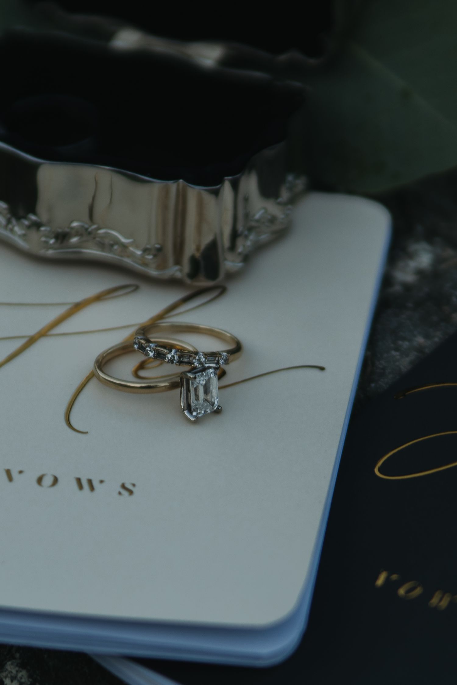 Diamond engagement ring and wedding bands placed on a white vow book with gold lettering.