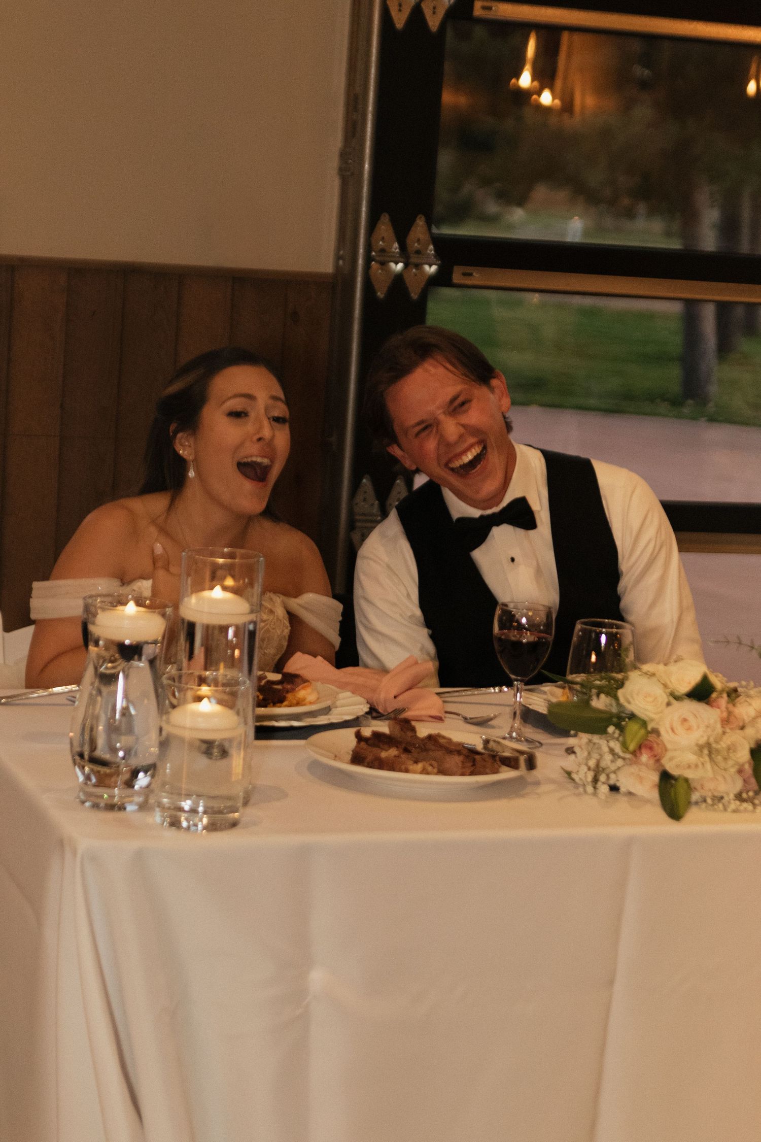 Newlyweds share laughter at their reception dinner table with candlelight.