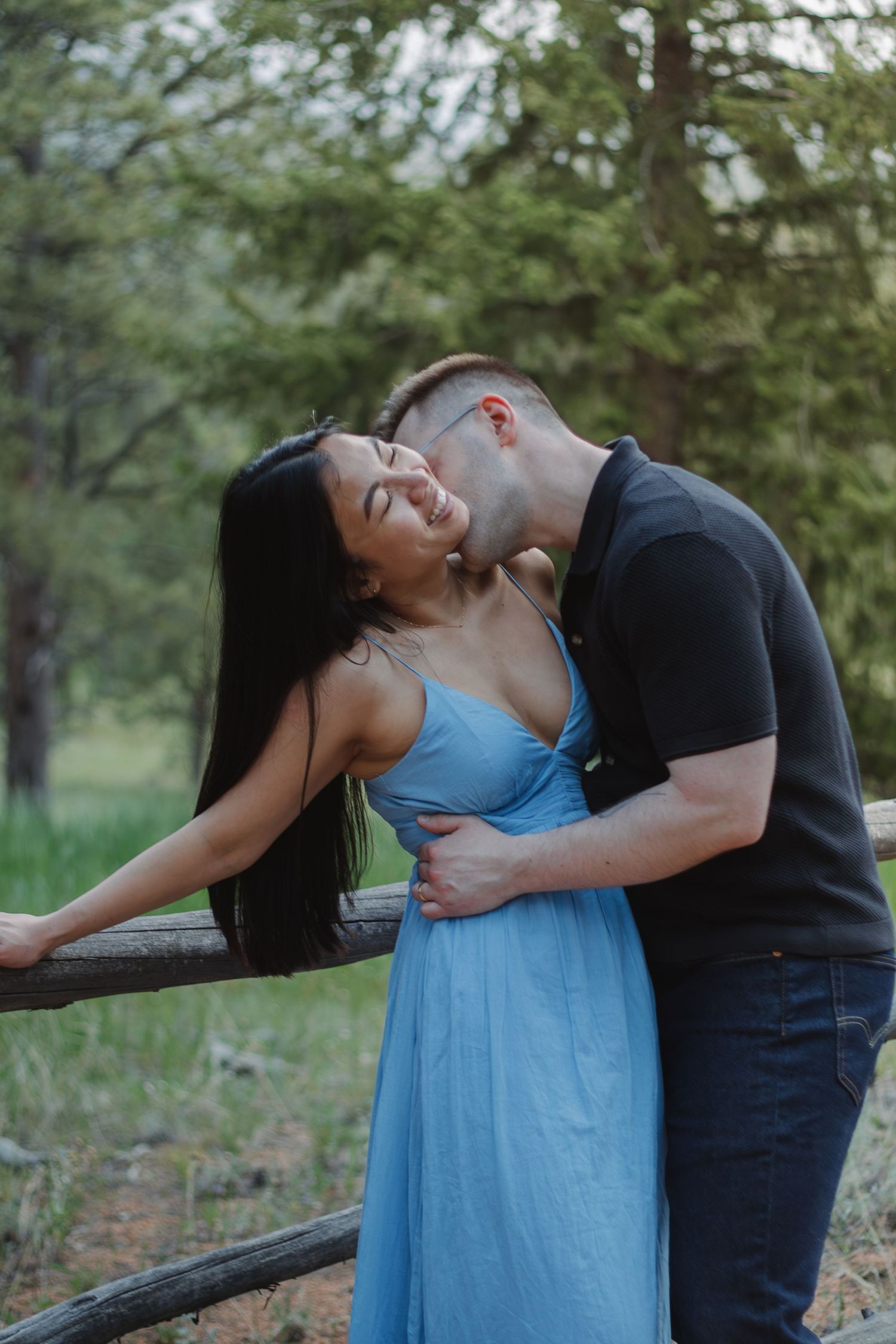 A couple shares an intimate moment by a rustic fence while wearing casual attire and a blue dress.