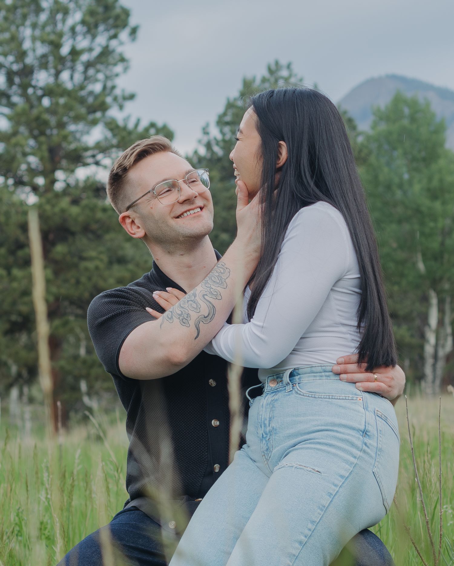 A joyful couple shares a playful moment in a mountain meadow with pine trees in the background.