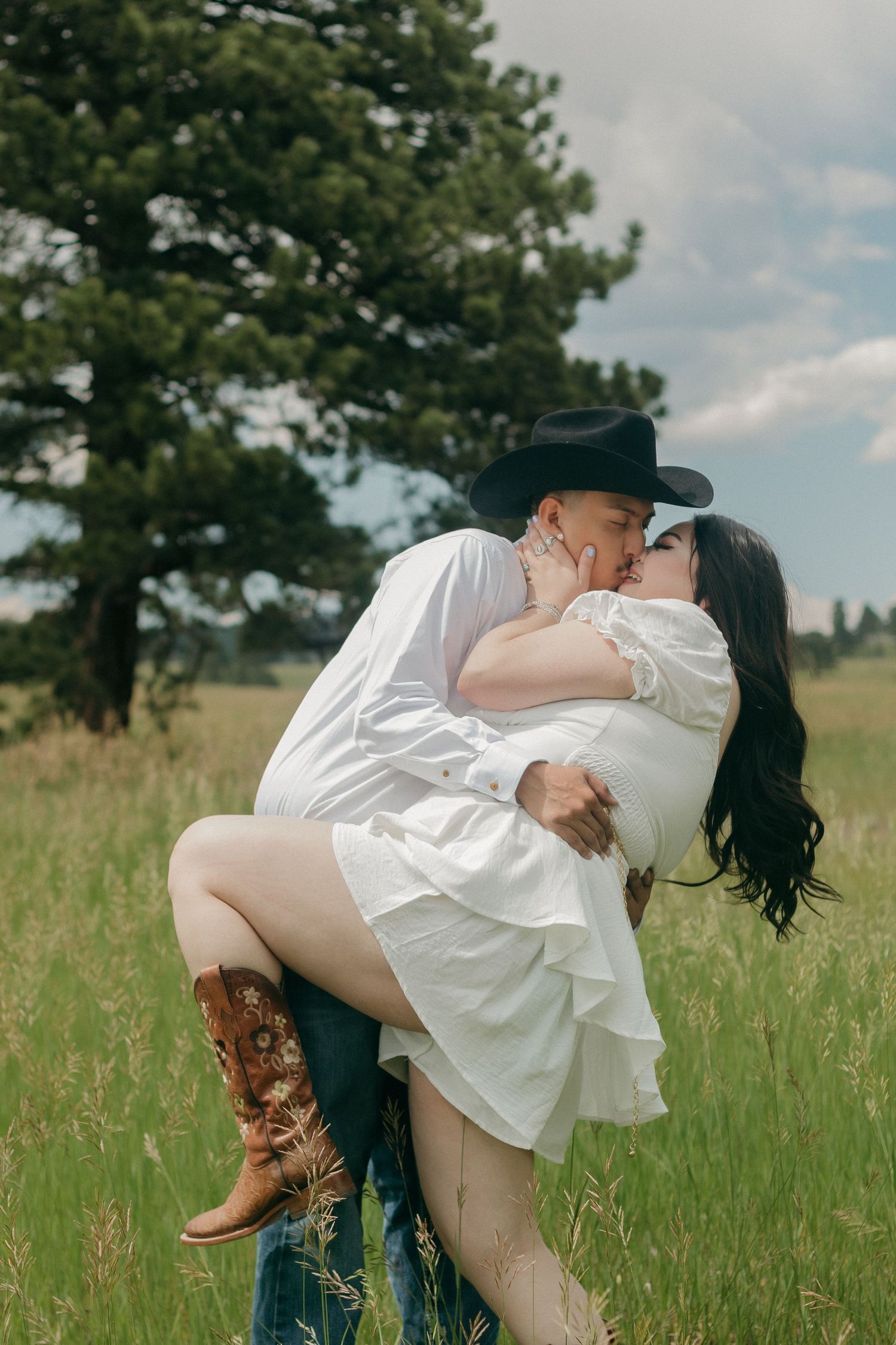 Romantic couple in western wear sharing tender moment in mountain meadow with pine trees.