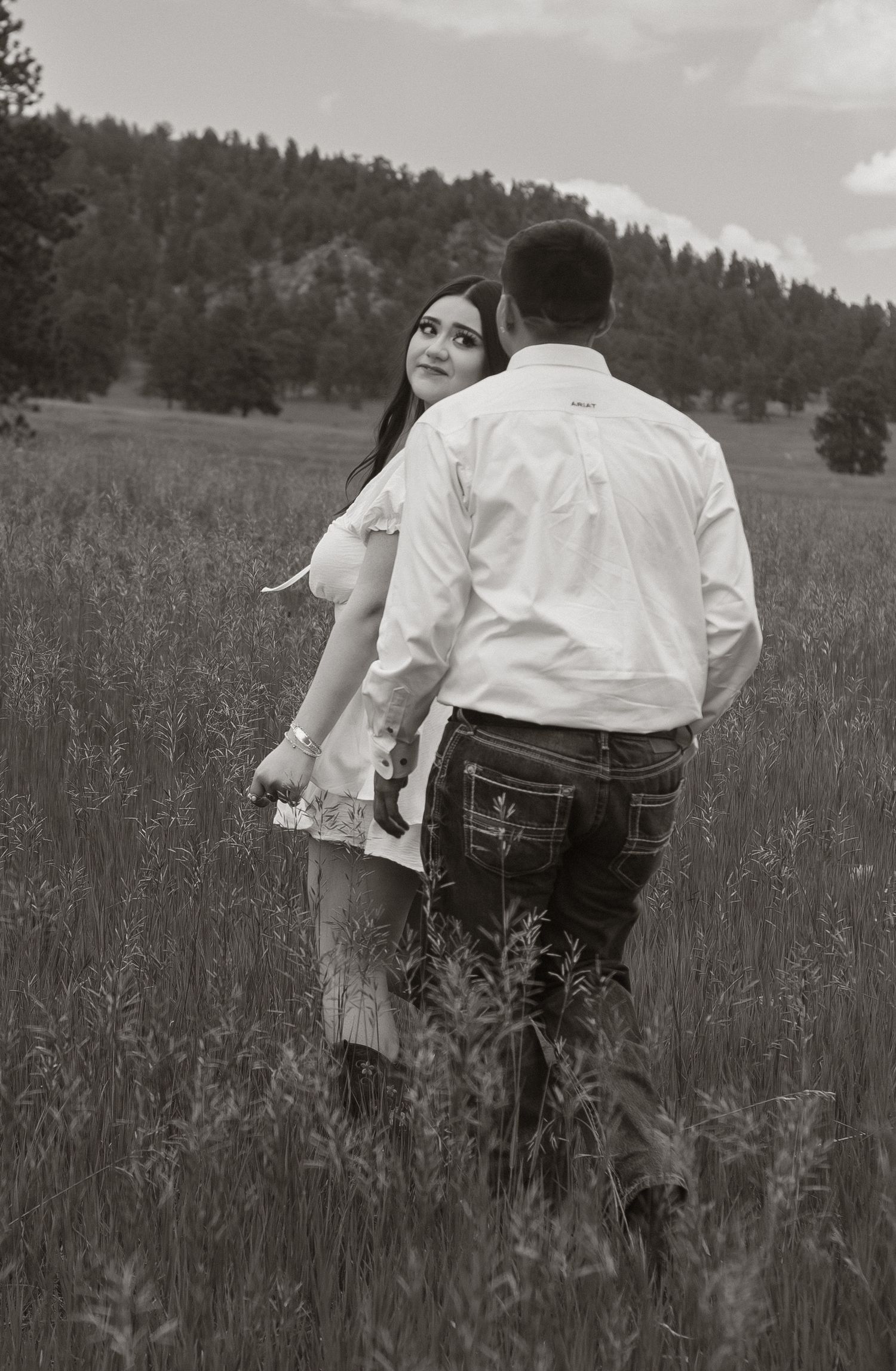 Black and white photo of a couple dancing together in a rustic field with trees in background.