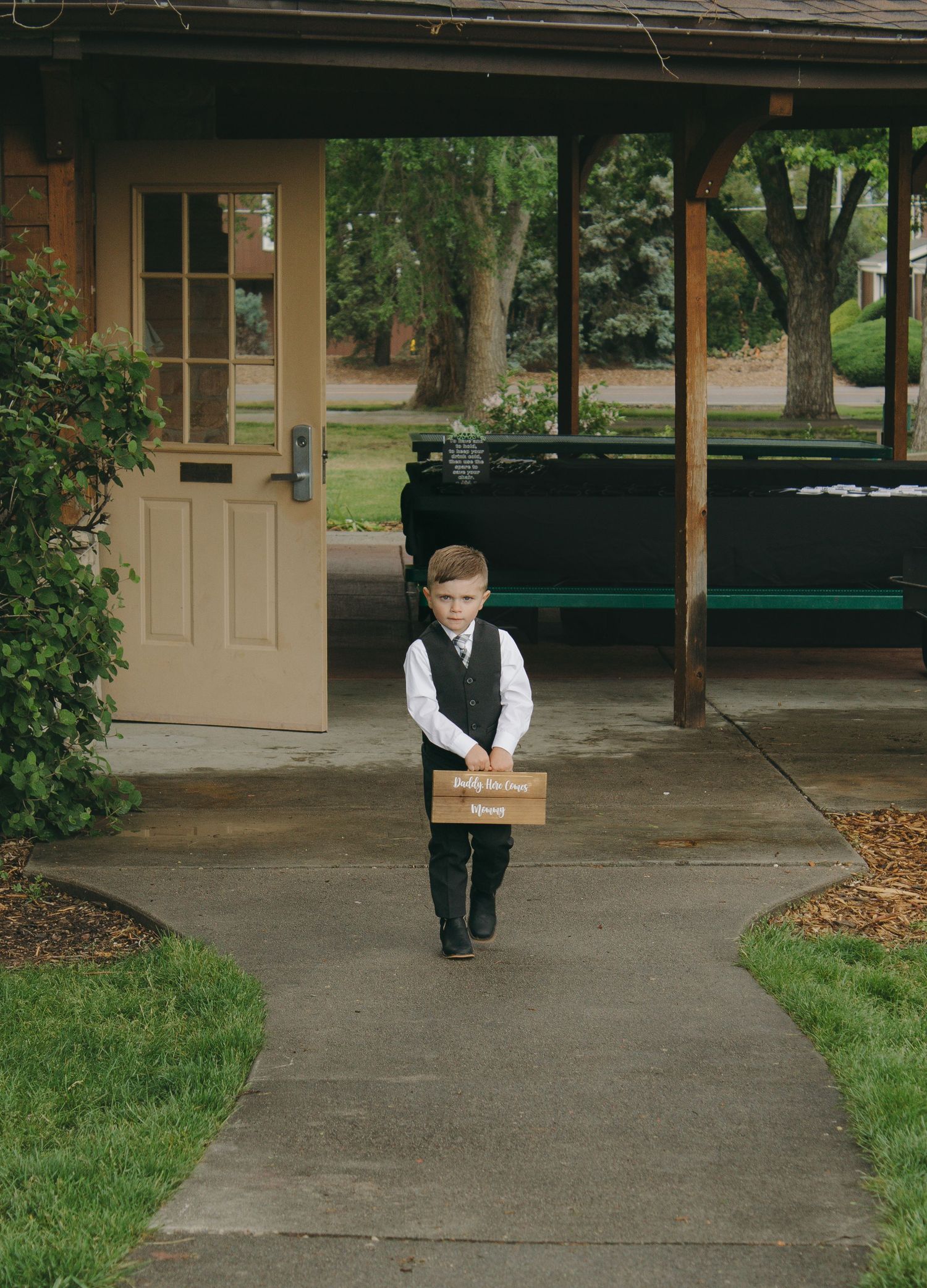 Young ring bearer in formal vest walks alone on a covered sidewalk.