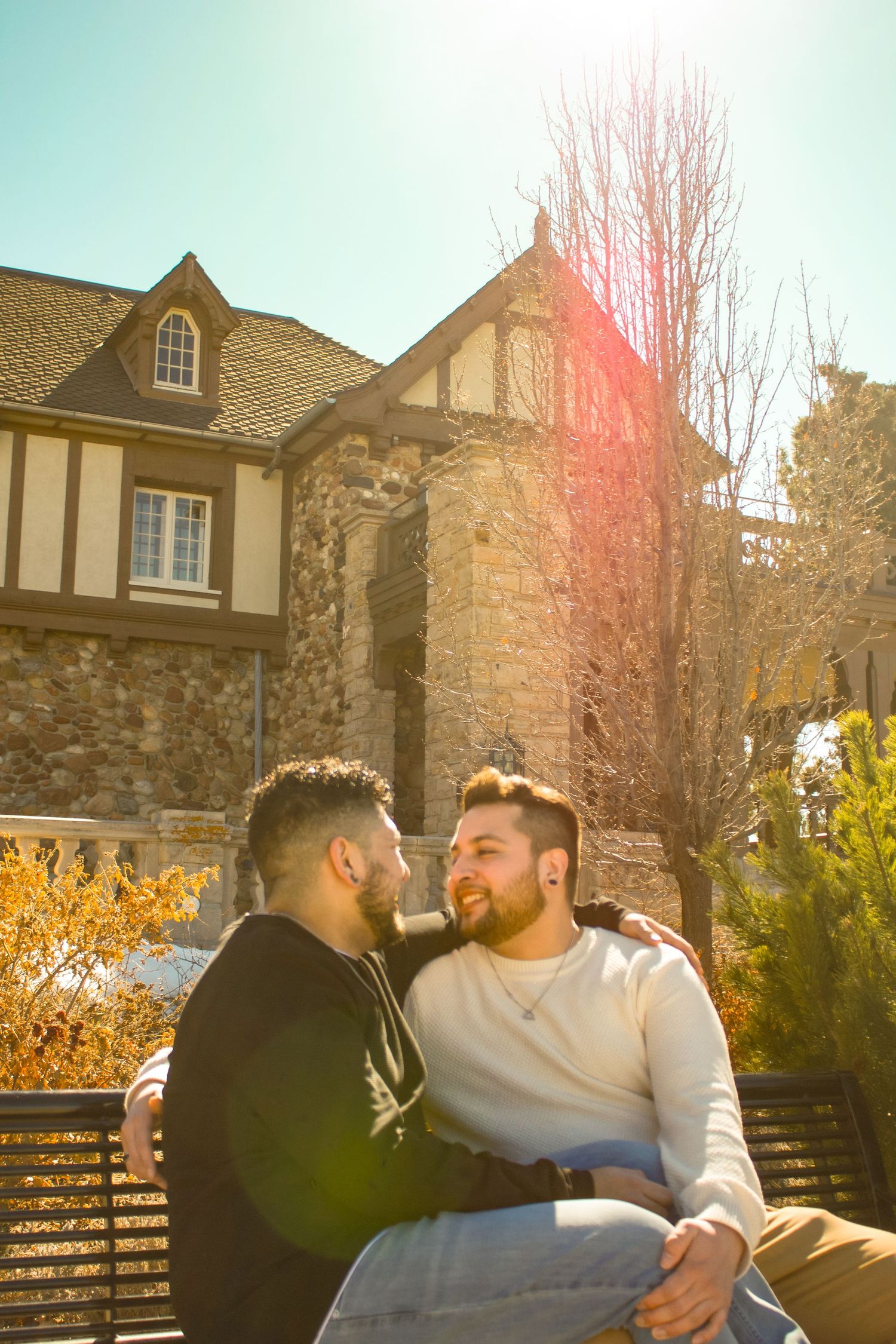 Two people sharing intimate moment on bench with stone buildings and autumn foliage in background.