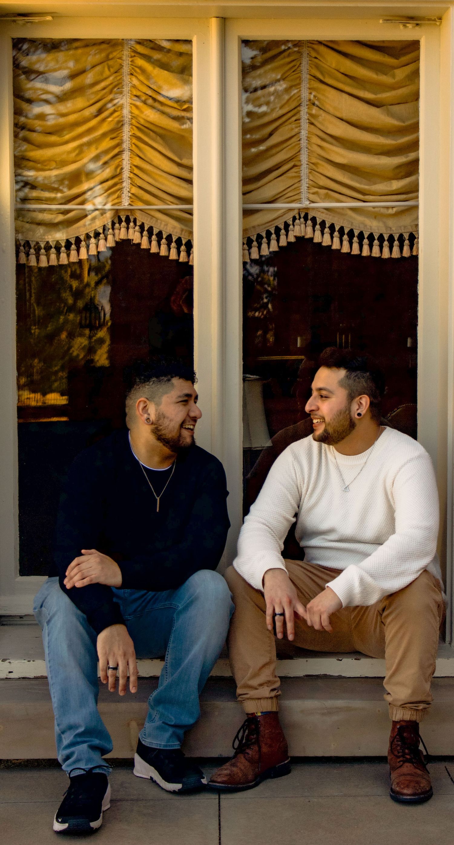 Two people sitting and chatting on porch steps with ornate window coverings behind them.