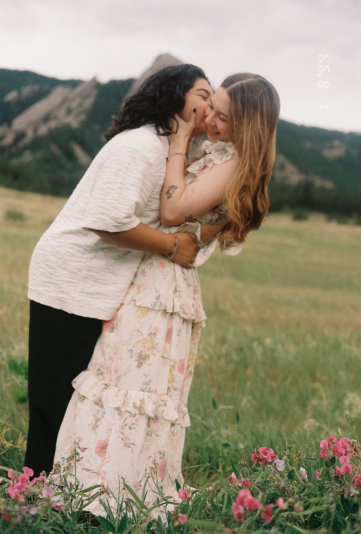 Couple sharing a romantic kiss in a wildflower meadow with mountain backdrop, one wearing a floral ruffle dress.