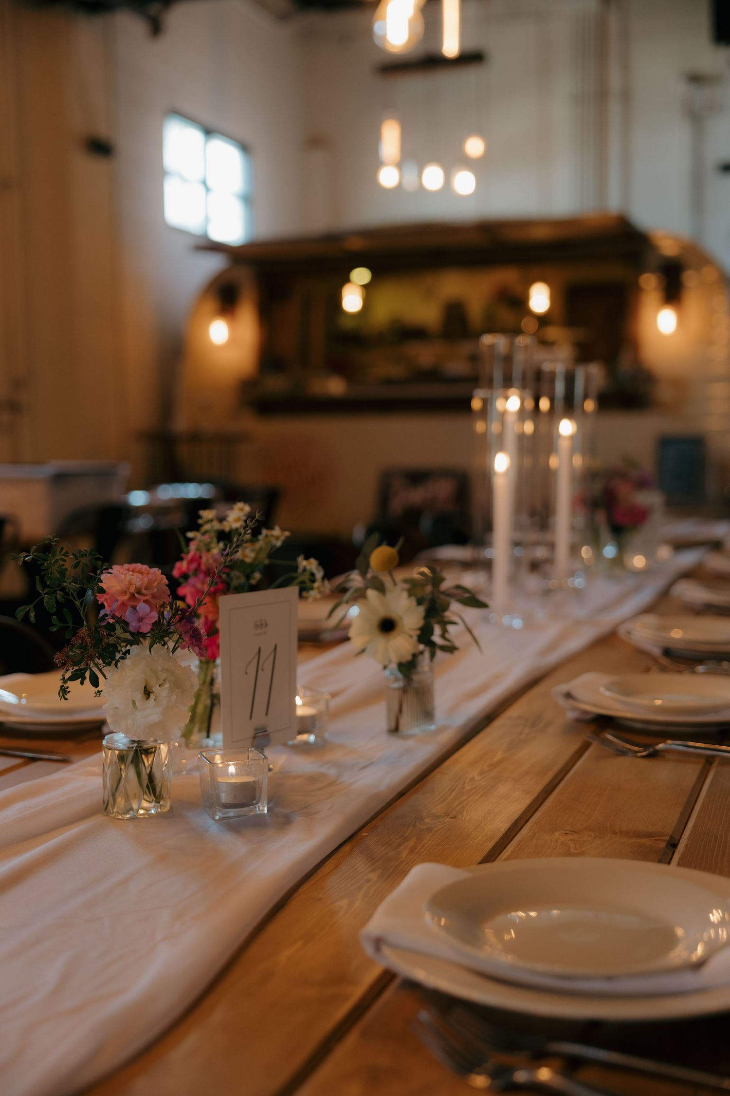 Elegant rustic wedding reception table with floral centerpieces, candles, and white plates on a wooden farm table.