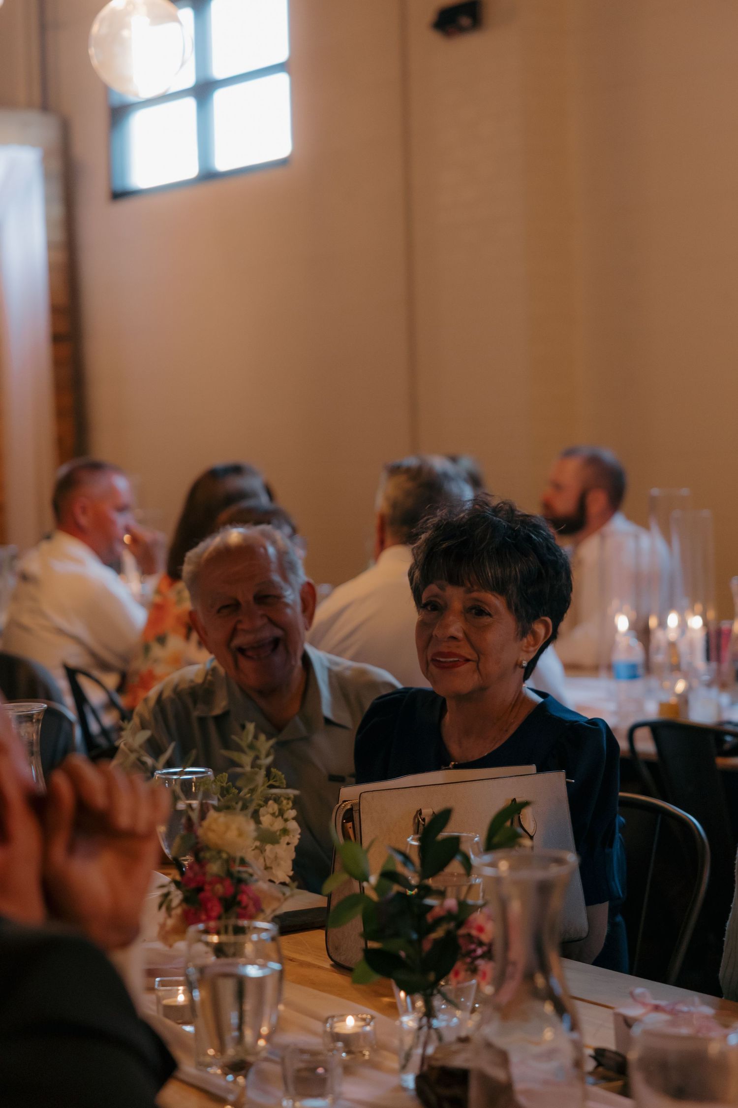 Smiling older couple enjoys wedding reception dinner at candlelit table decorated with fresh floral centerpieces.