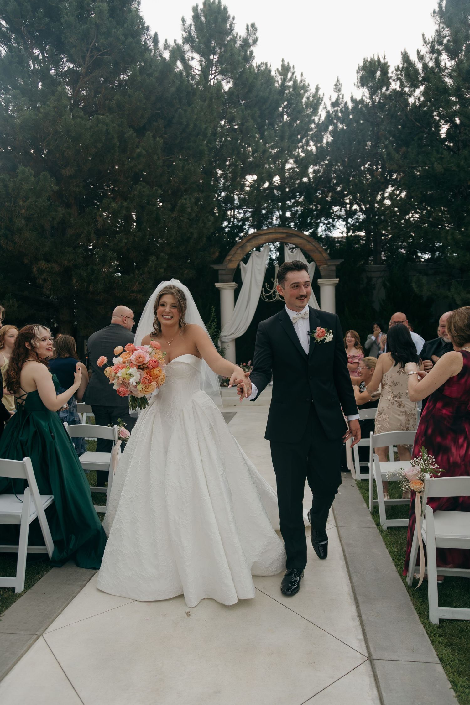Bride and groom walk down outdoor wedding aisle, bride holding colorful bouquet, guests seated on white chairs.