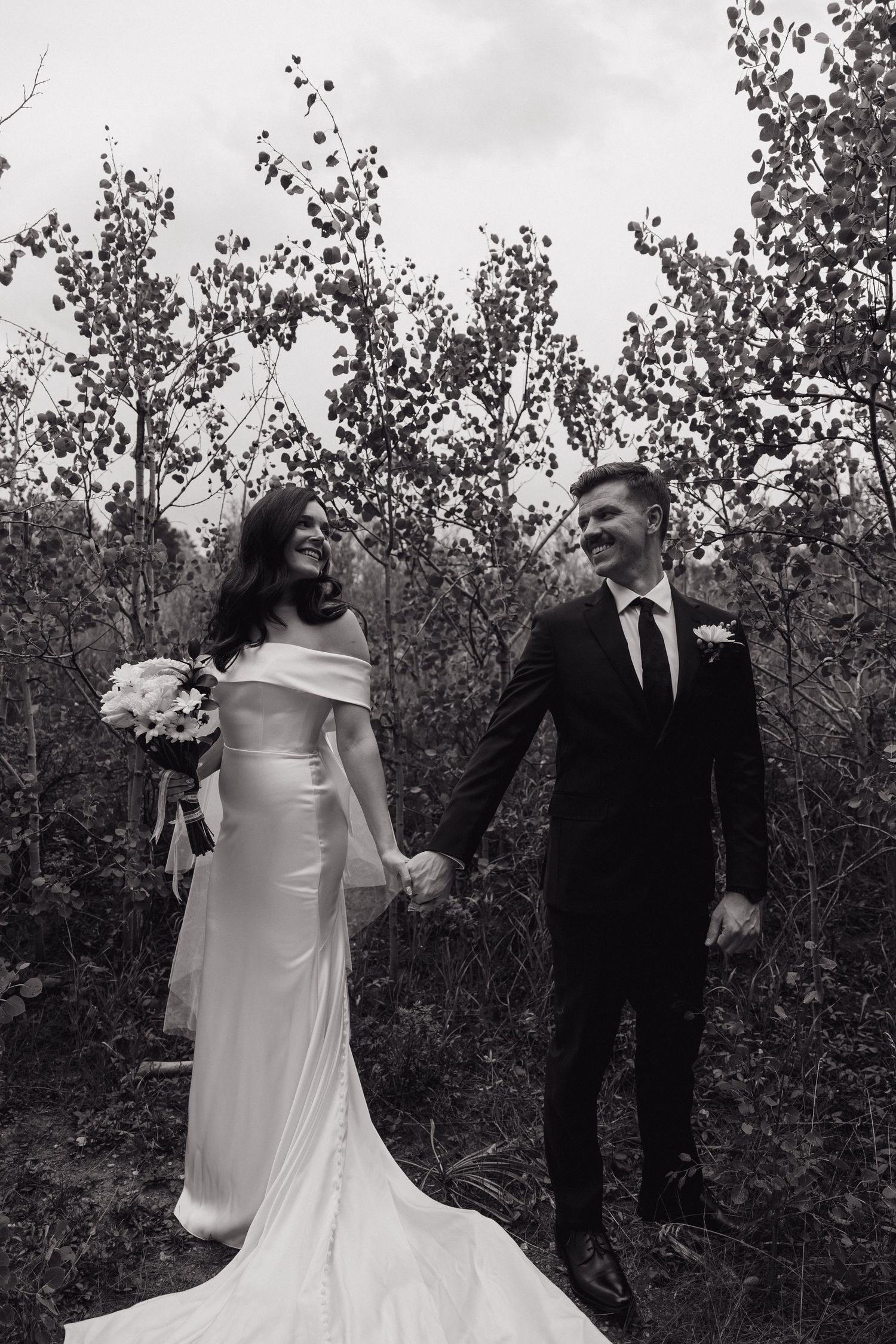 Black and white photo of smiling bride and groom holding hands walking through trees on their wedding day.
