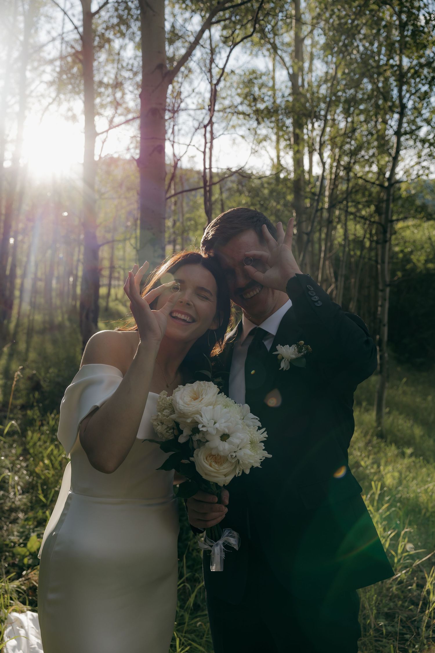 Joyful bride and groom pose playfully in sunlit aspen forest with white wedding bouquet.