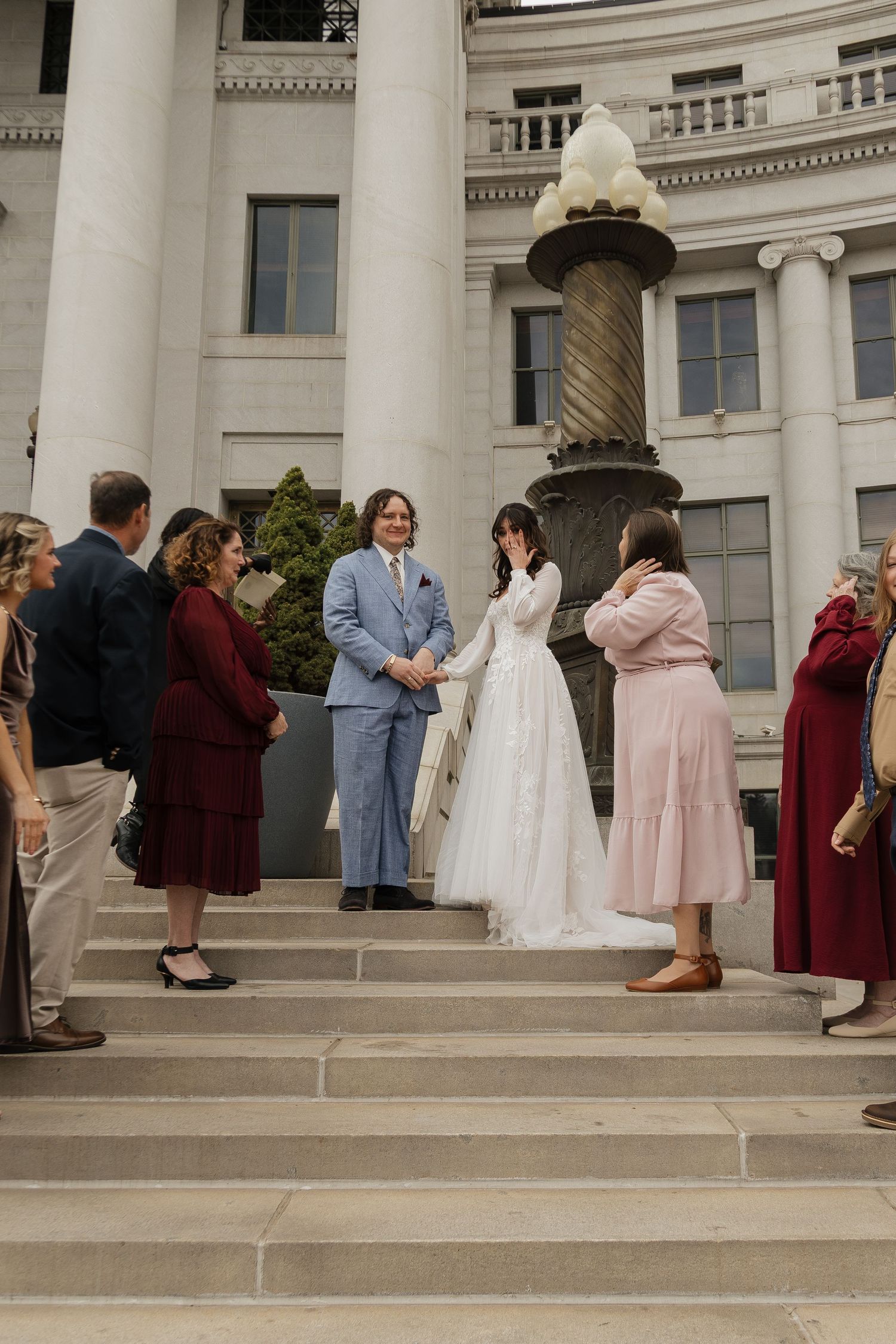 A wedding ceremony on grand stone steps with guests in burgundy dresses and a couple in light blue and white attire.