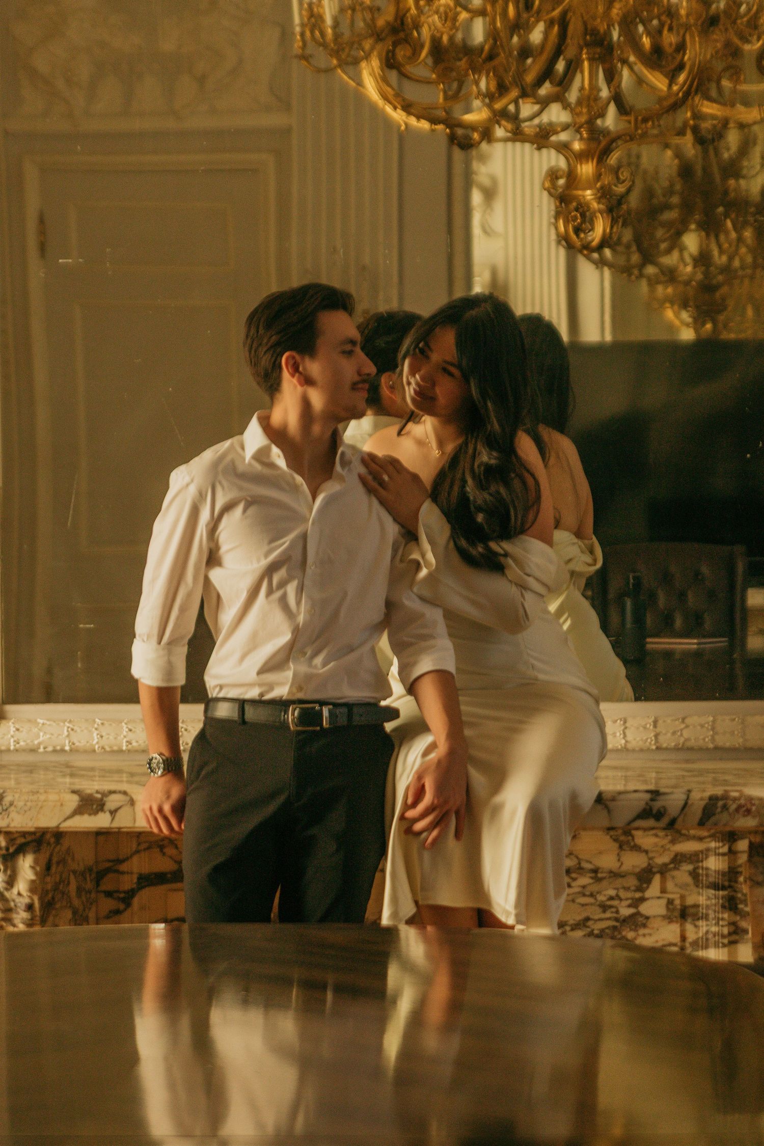Romantic couple poses beneath golden chandelier in elegant vintage room.