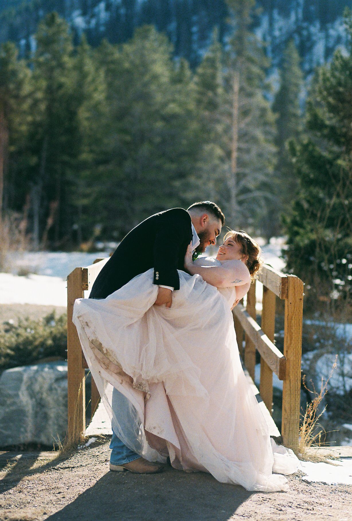 A romantic winter wedding couple shares intimate dips and kisses on a wooden bridge in a snowy mountain setting.