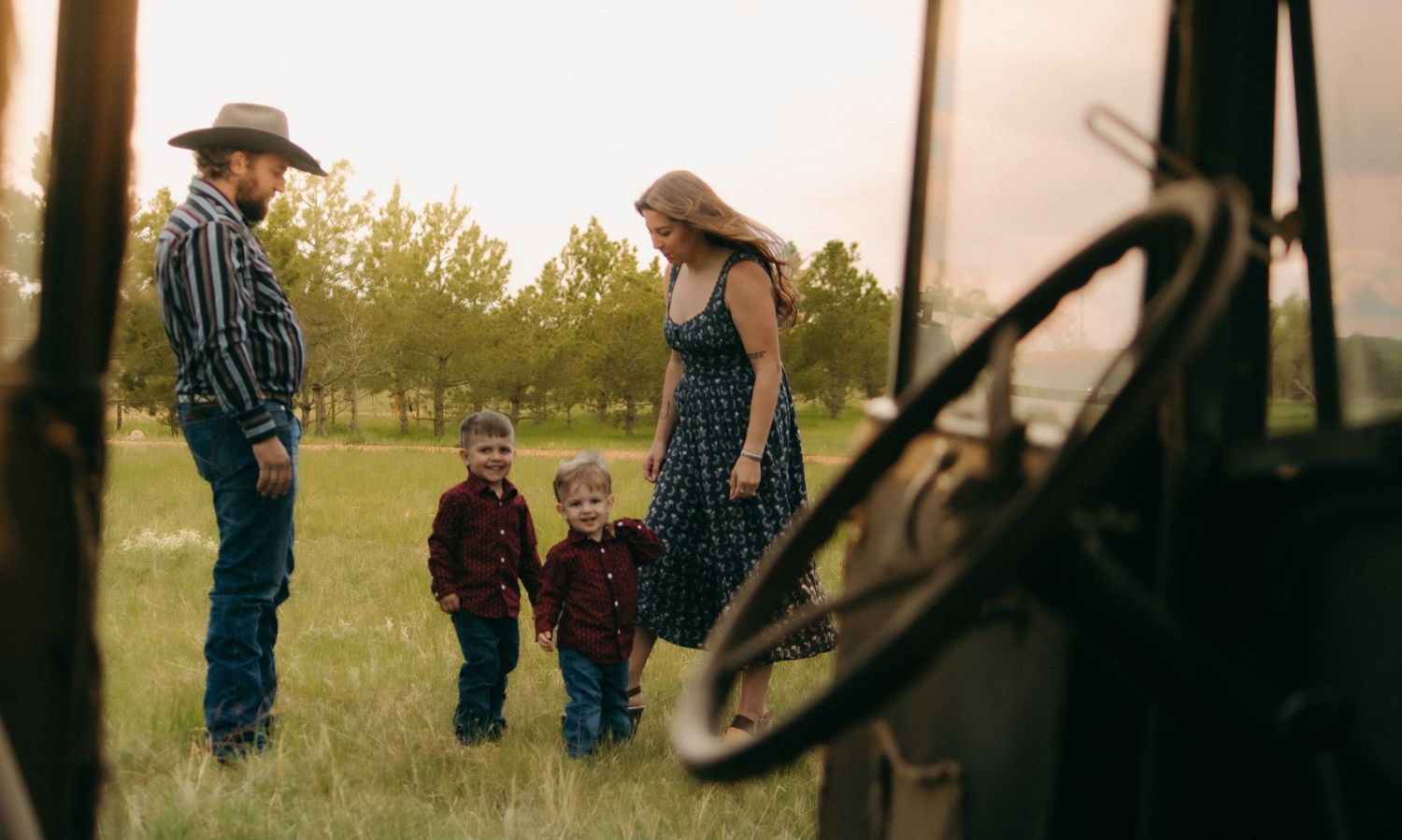 A family enjoys a tender moment together in a grassy field at sunset with a rustic fence in the background.