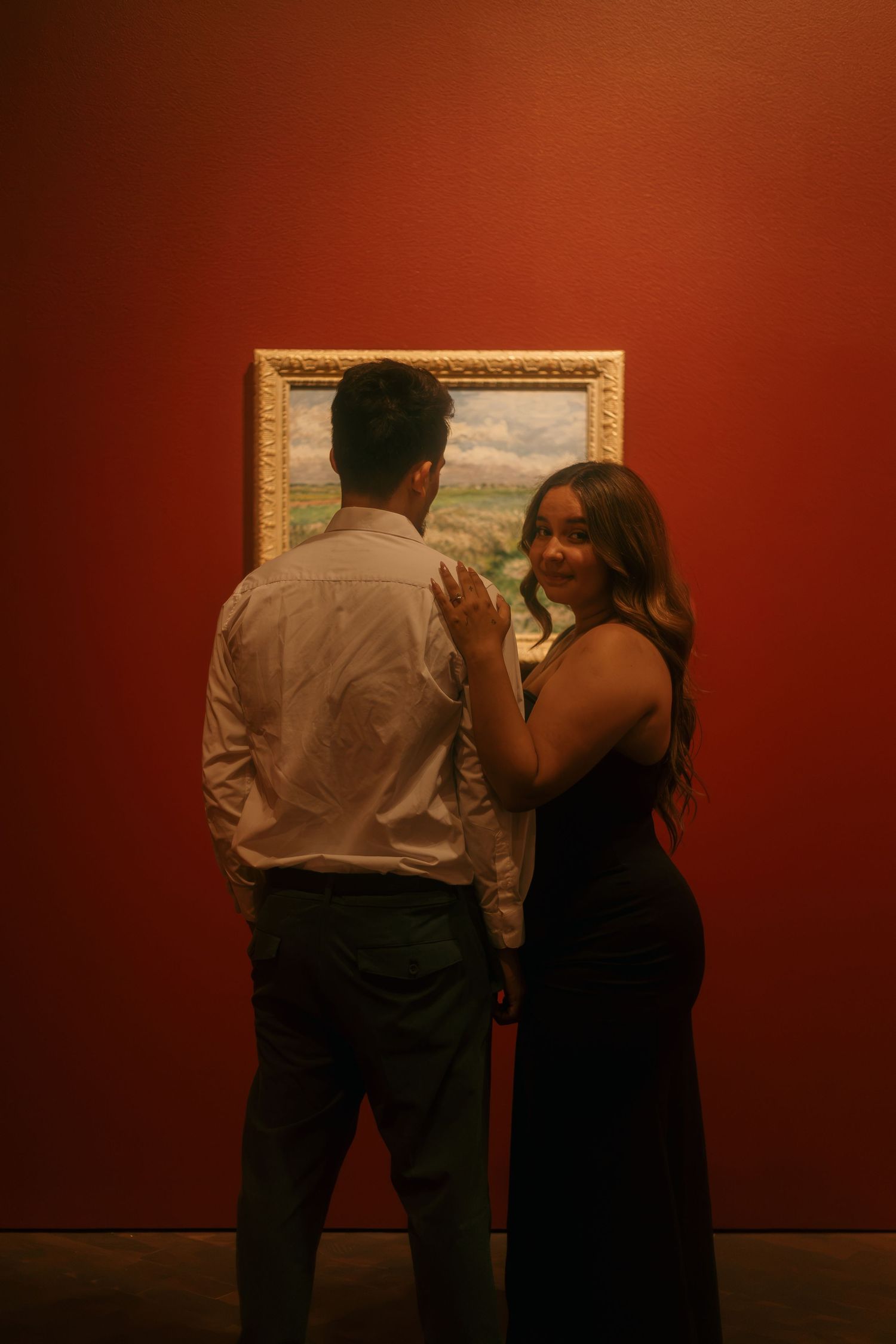 Couple standing before a framed landscape painting on a deep red gallery wall.
