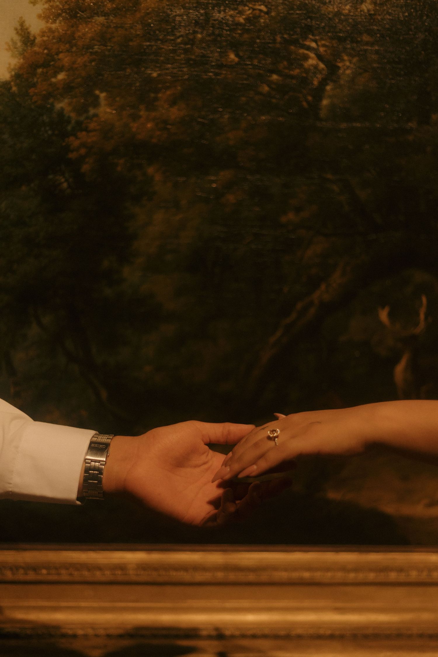 Two hands reaching toward each other with an engagement ring before a classical landscape oil painting.