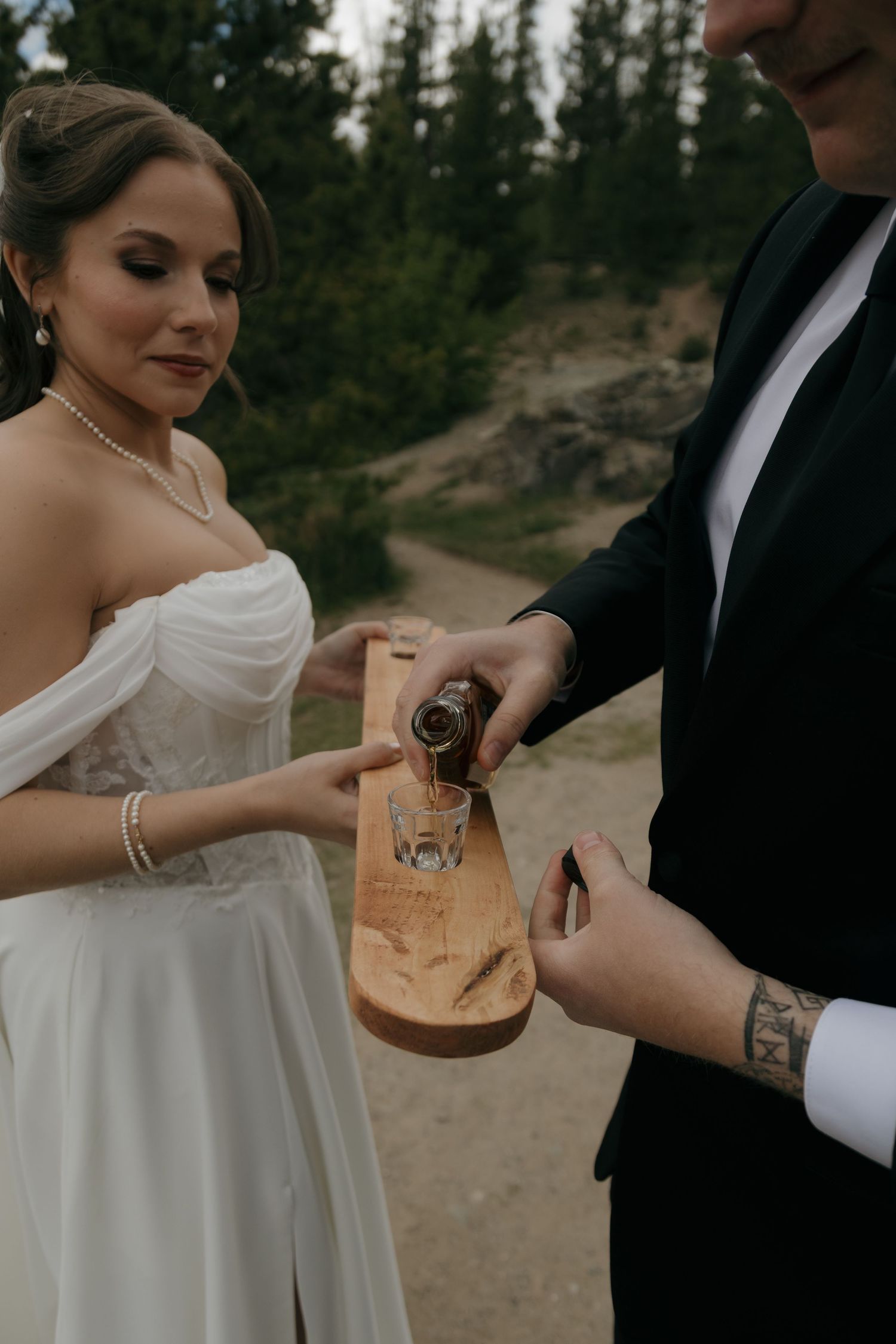 Bride and groom perform a whiskey unity ceremony, pouring bourbon into glasses on a rustic wooden board outdoors.