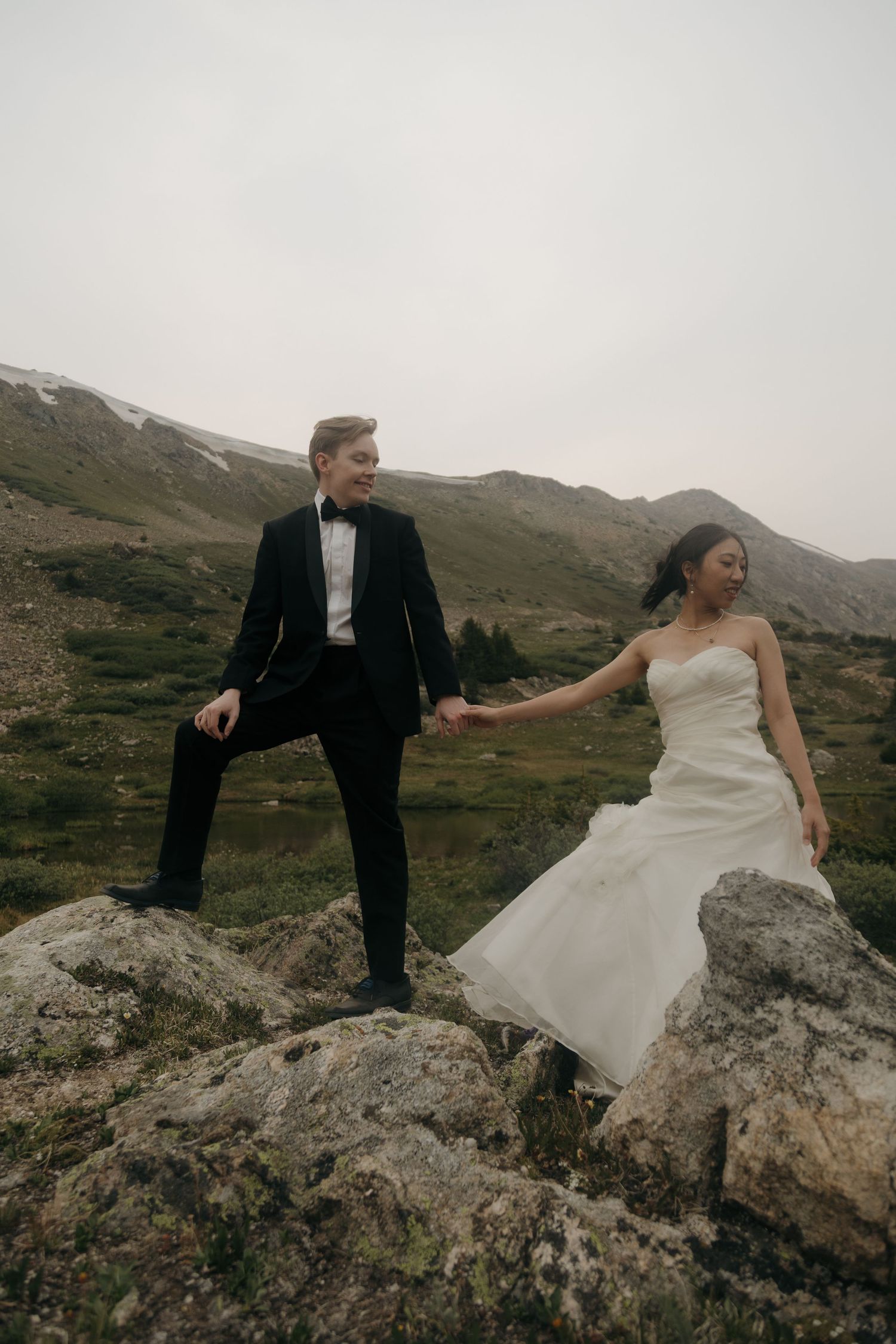 Groom helping bride climb rocky terrain while holding hands during their mountain elopement adventure.