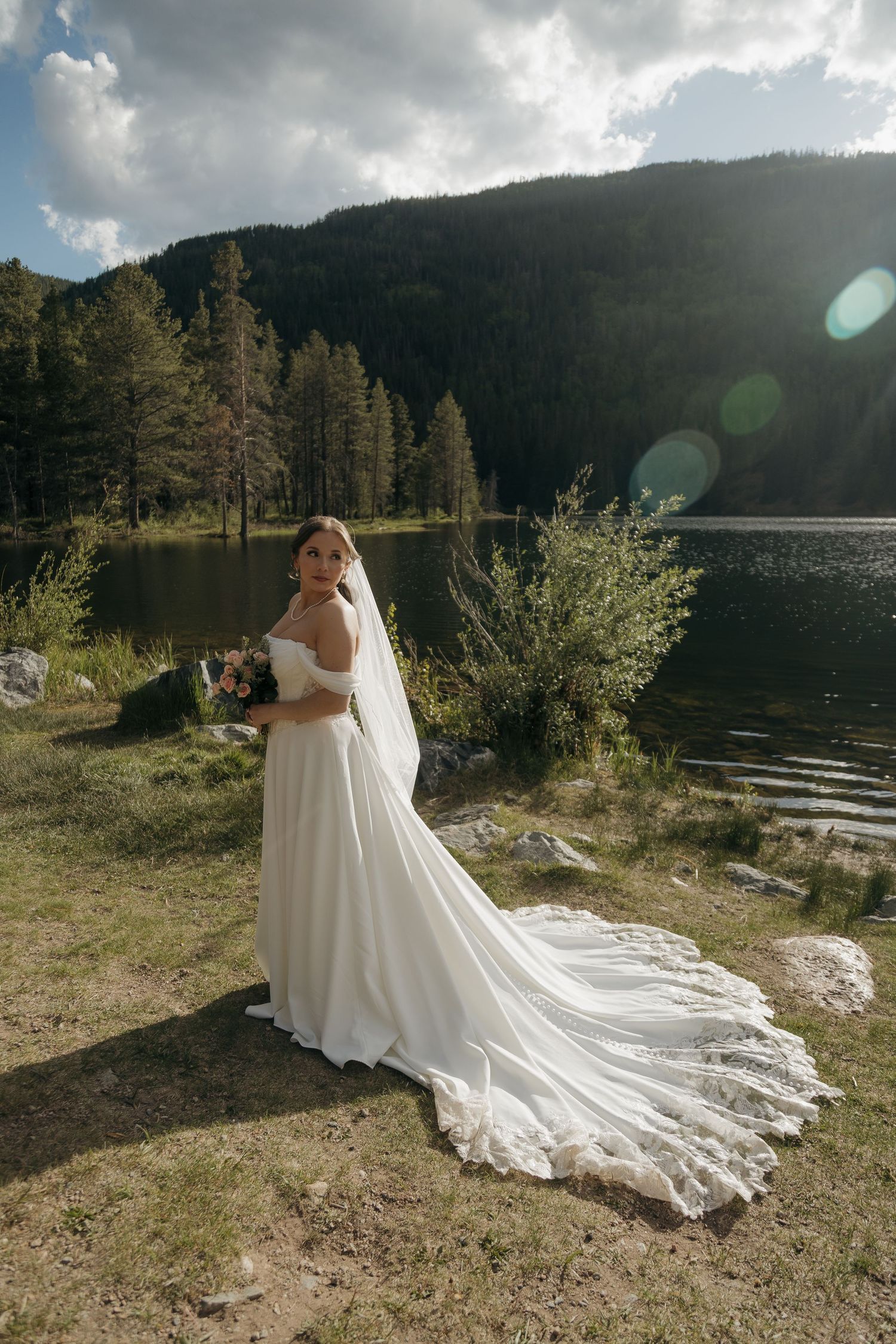 Bride in elegant white gown with lace train posing by a sunlit mountain lake.