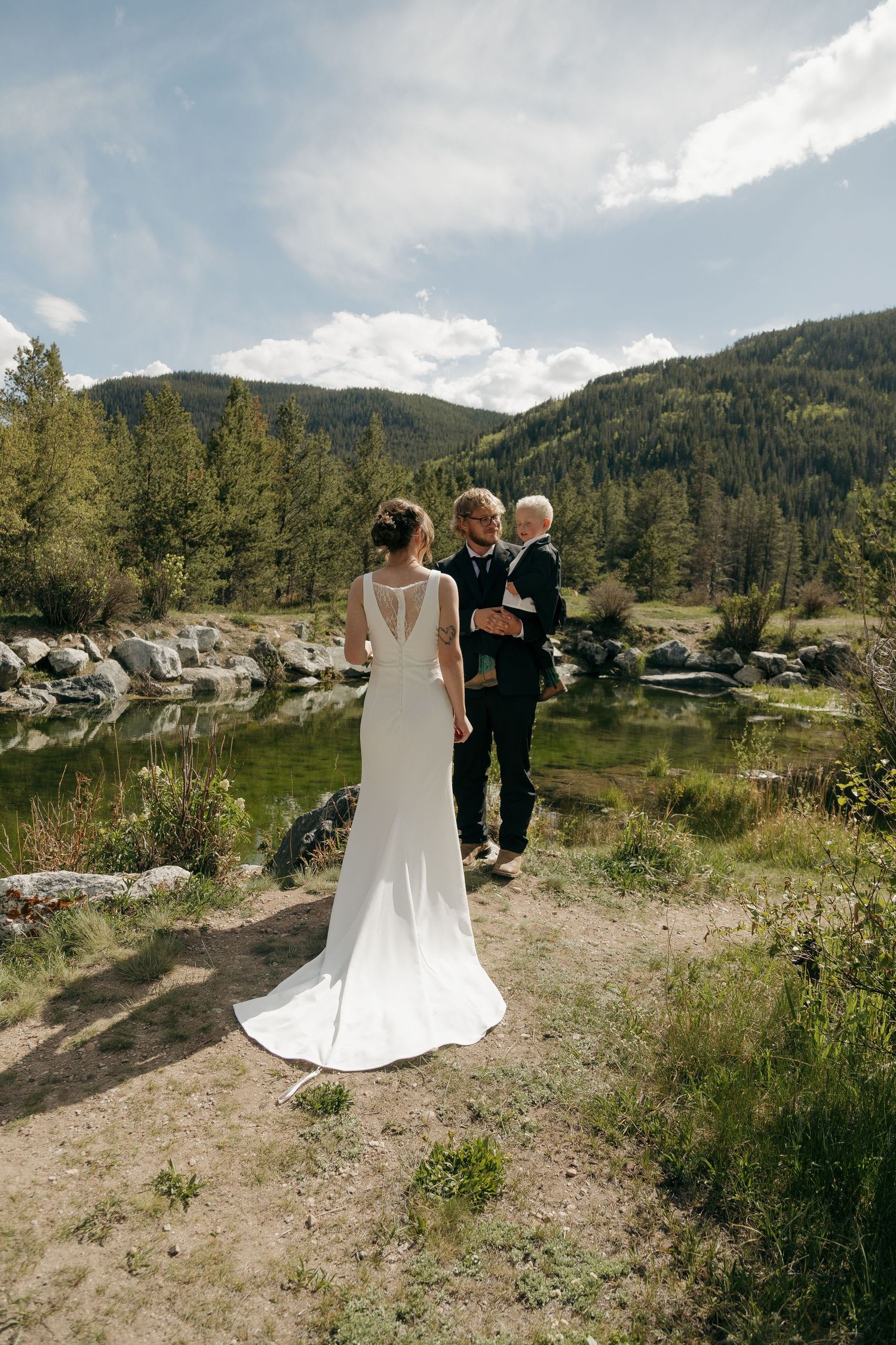 Bride in white gown and groom in black suit stand by a mountain lake surrounded by pine trees.