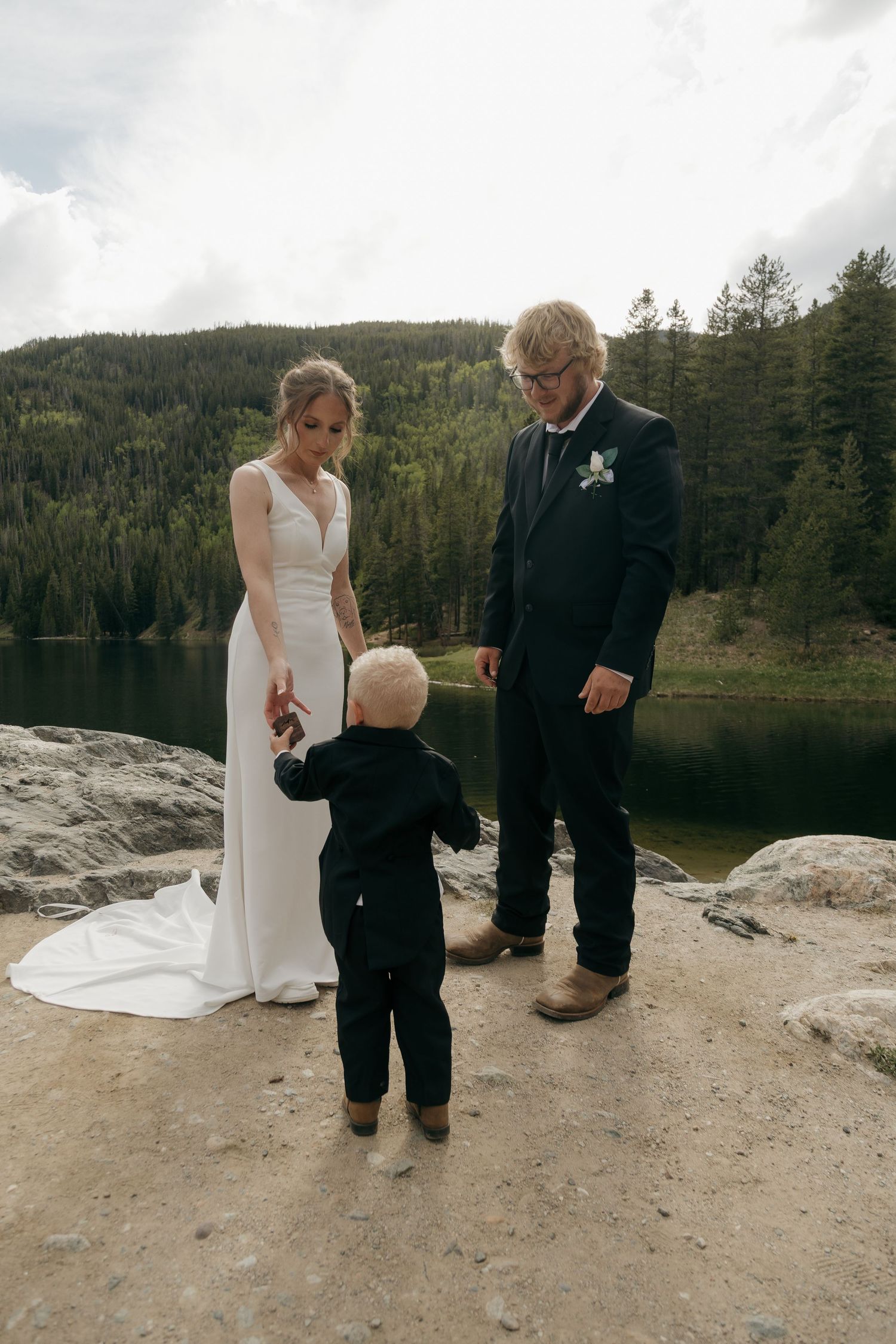 A bride and groom in wedding attire stand with a young child near a scenic mountain lake.