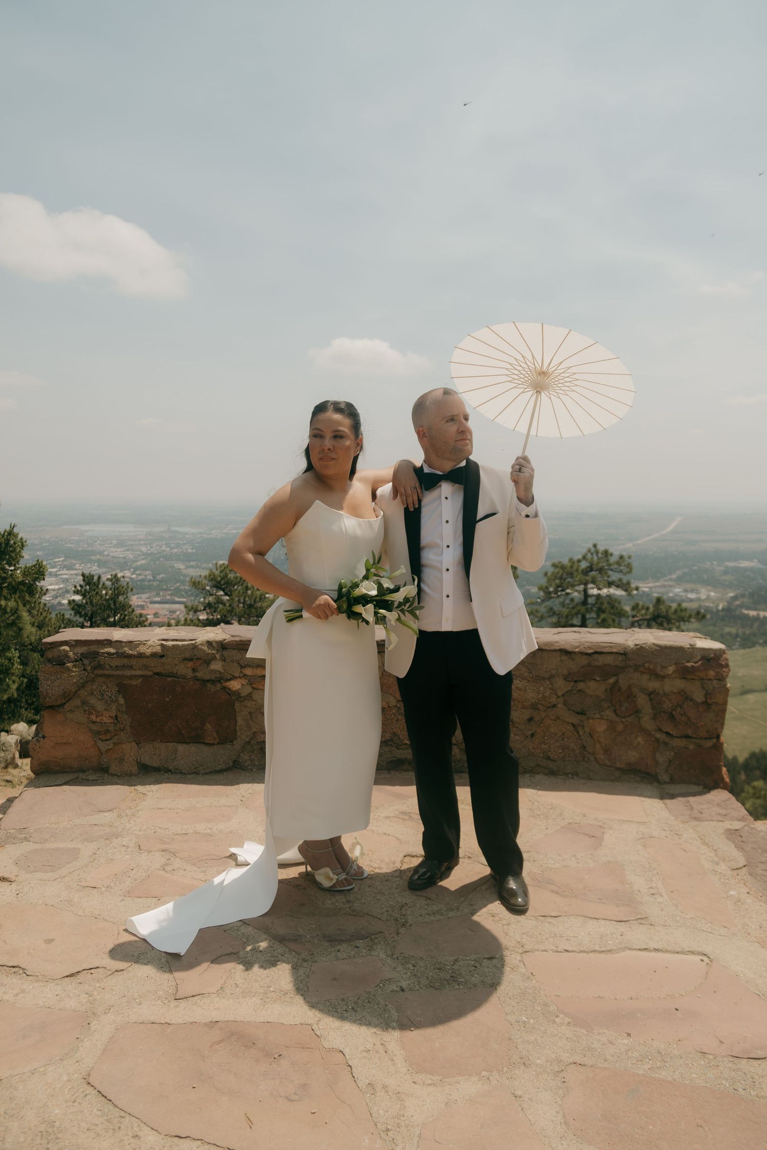 Bride and groom pose on a stone terrace with panoramic views, she holds flowers while he raises a white parasol.