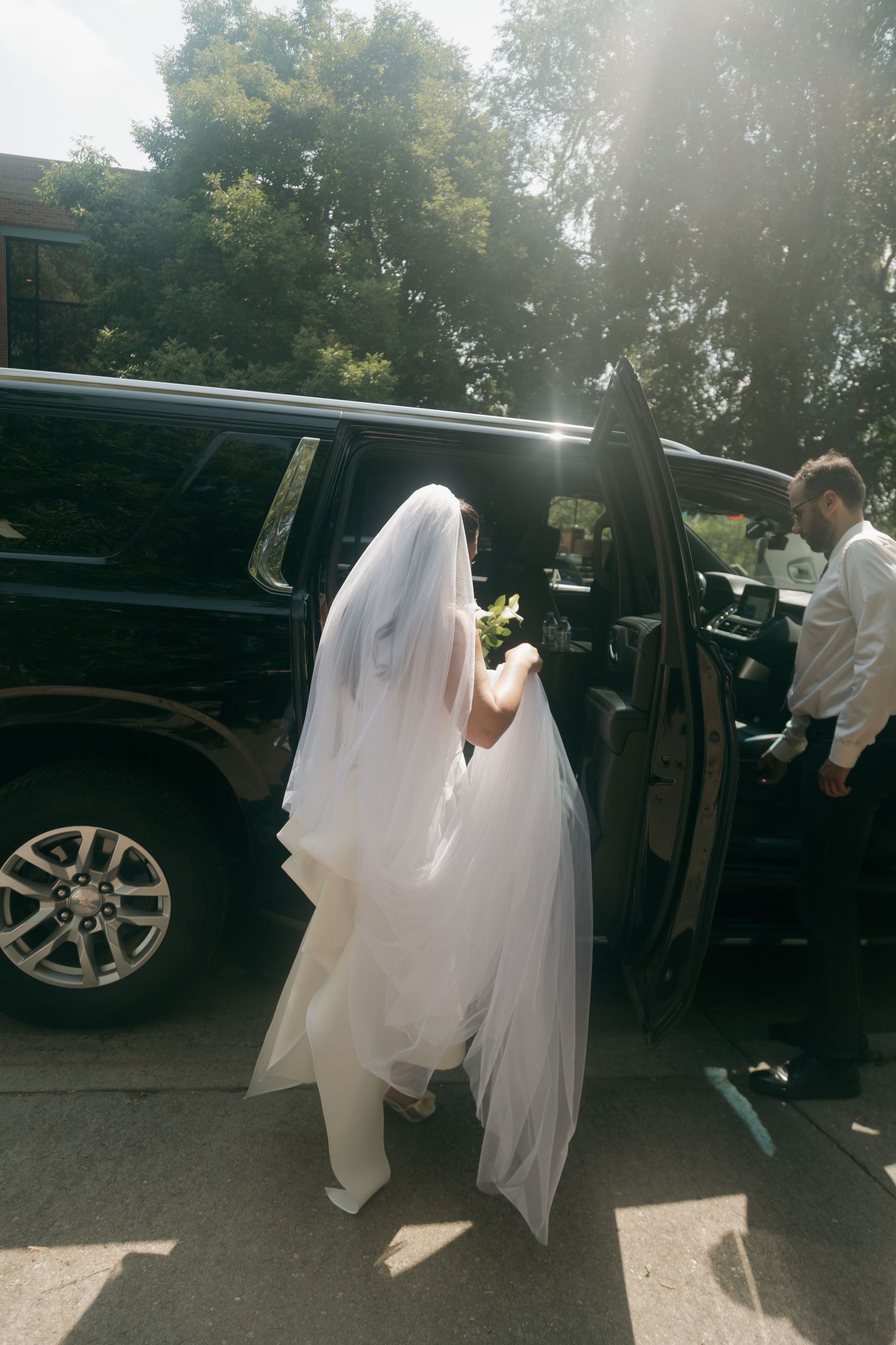 Bride in white veil and gown entering black SUV on sunny wedding day.